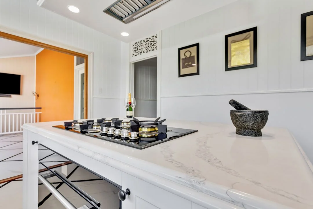 White kitchen island with a black gas stove, black framed pictures on the wall, grey mortar and pestle, champagne bottle and glasses, with a doorway leading to living room with orange wall and a mounted TV.