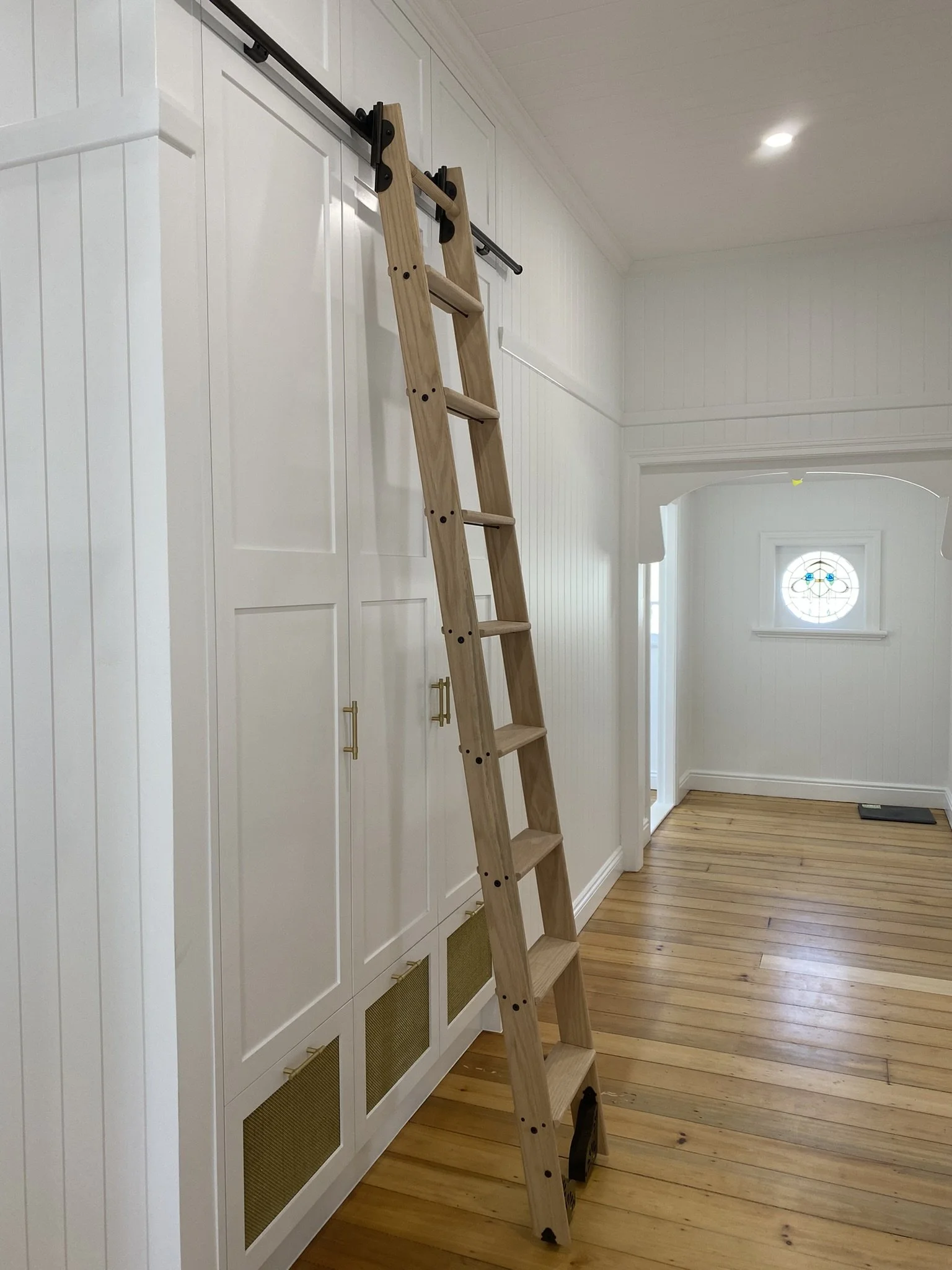 Wooden ladder leaning against a white cabinet in a bright room with wooden flooring and a small stained glass window.