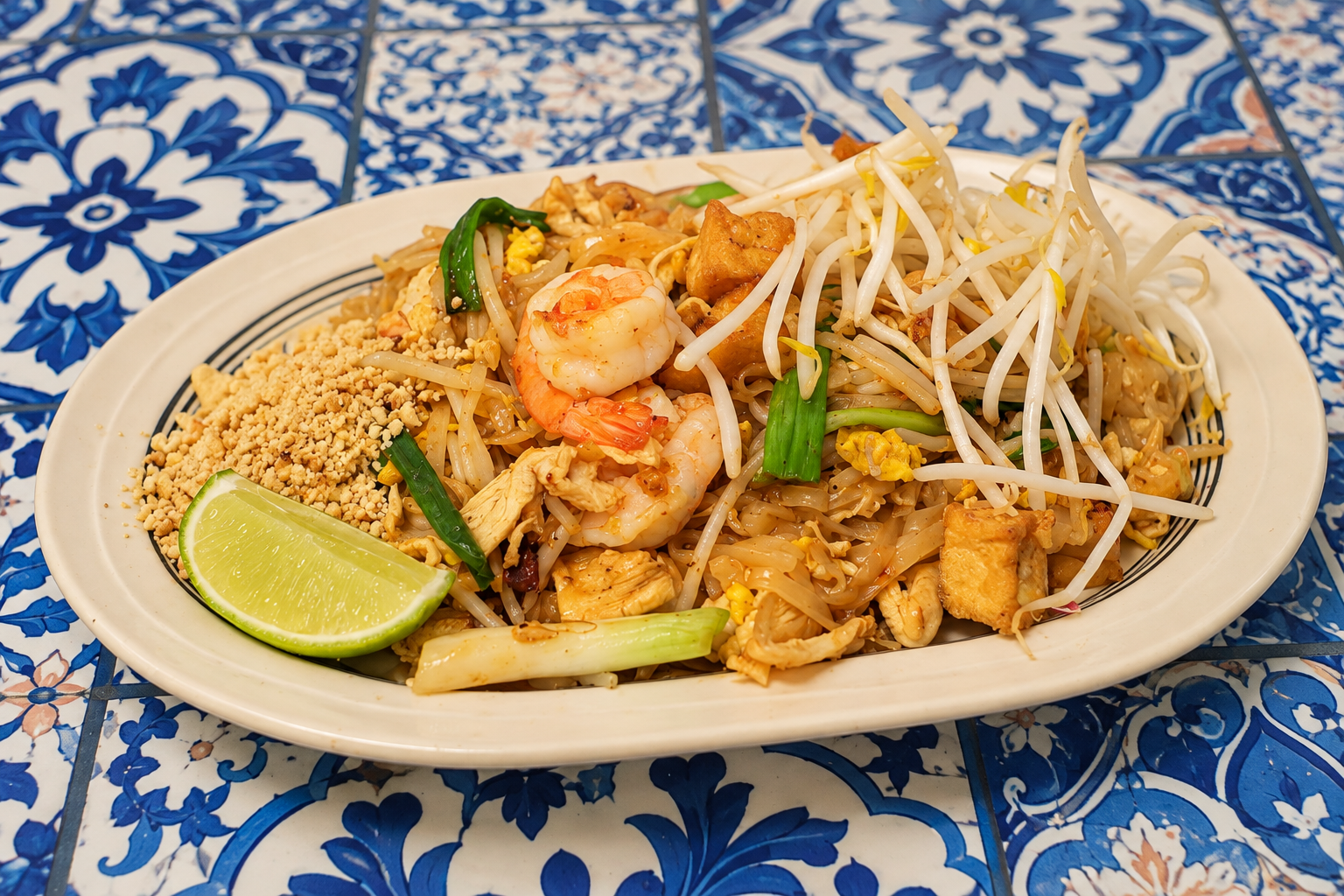Plate of Pad Thai with shrimp, bean sprouts, green onions, crushed peanuts, and a lime wedge on a blue and white patterned table.