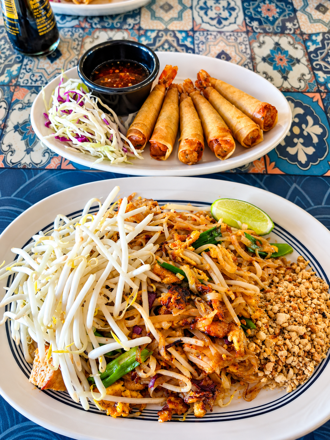 Plate of Pad Thai with bean sprouts, chopped peanuts, and a lime wedge; another plate with spring rolls, a dipping sauce, and shredded cabbage with purple accents.