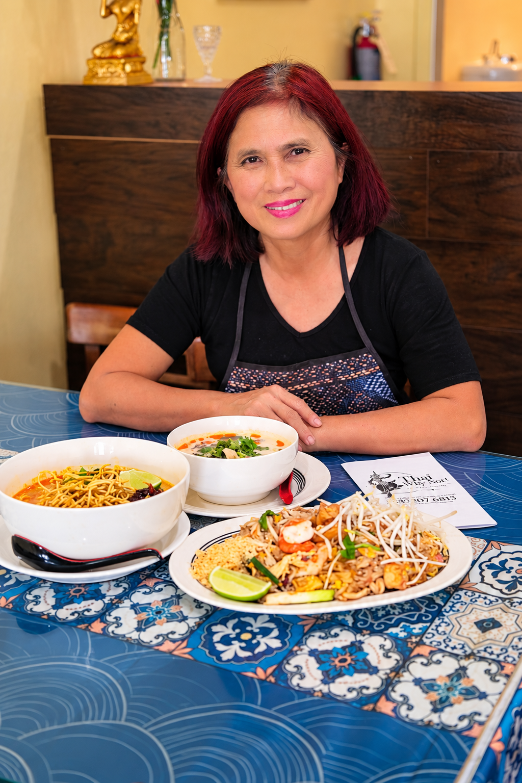 A woman with red hair smiling at a table with various Thai dishes, including noodles, soup, and a seafood salad, in a restaurant setting.