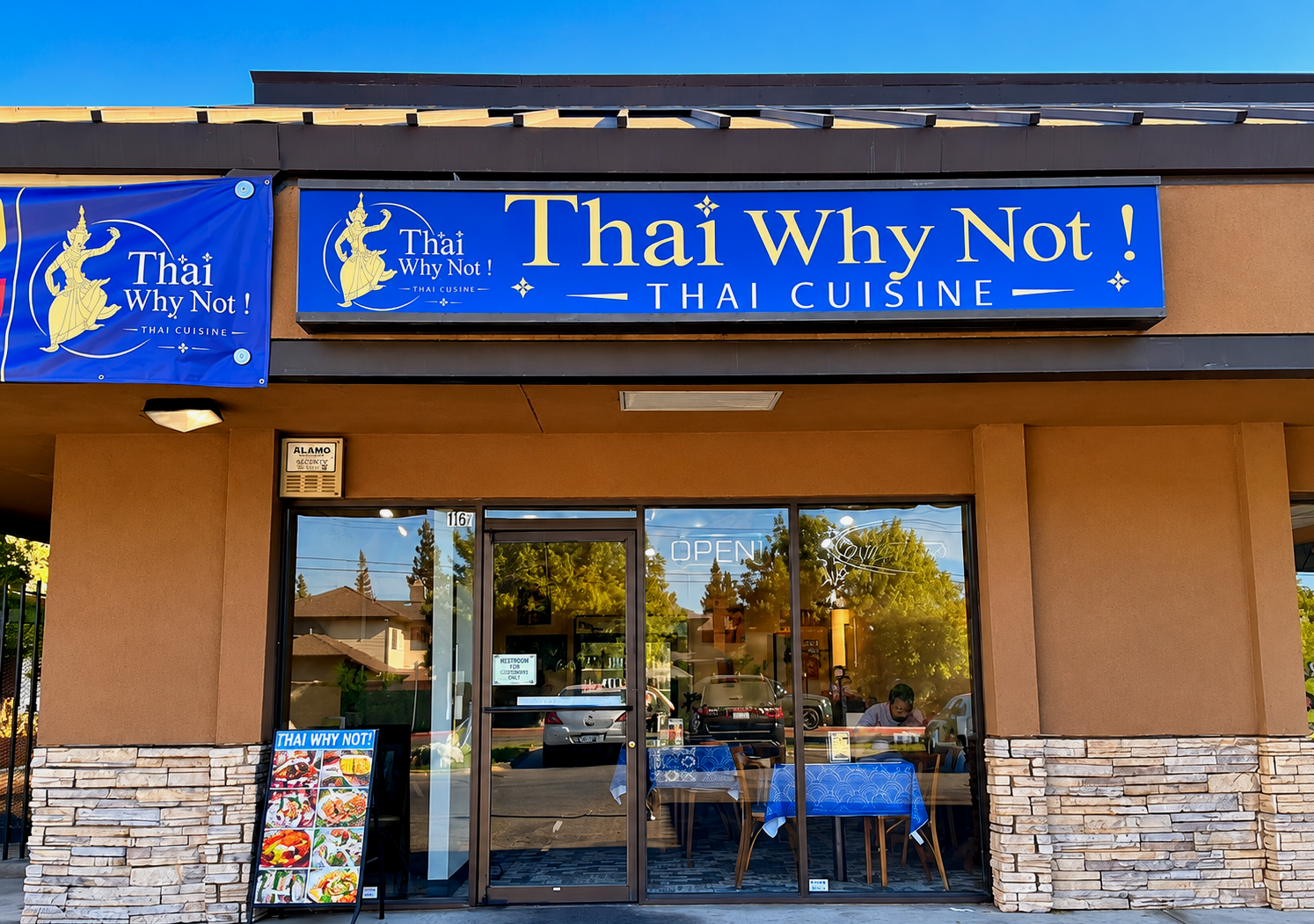 Exterior view of a Thai restaurant called 'Thai Why Not!' with a blue sign, glass doors, and outdoor seating. The restaurant has a neon 'Open' sign in the window, and a menu board featuring Thai dishes outside.