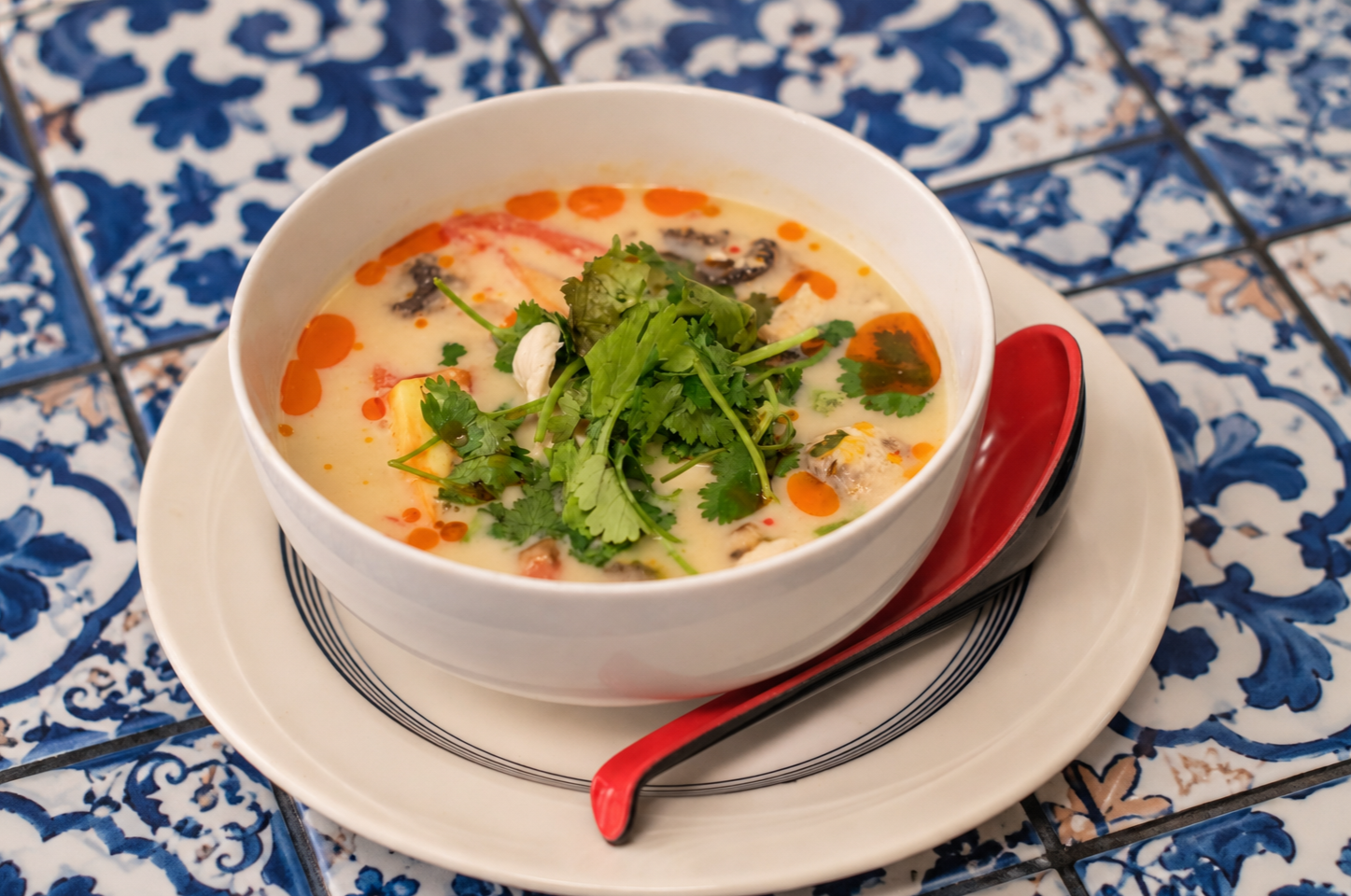 A bowl of Thai green curry chicken topped with cilantro and served with a red and black spoon on a white plate, placed on a blue and white patterned tile surface.