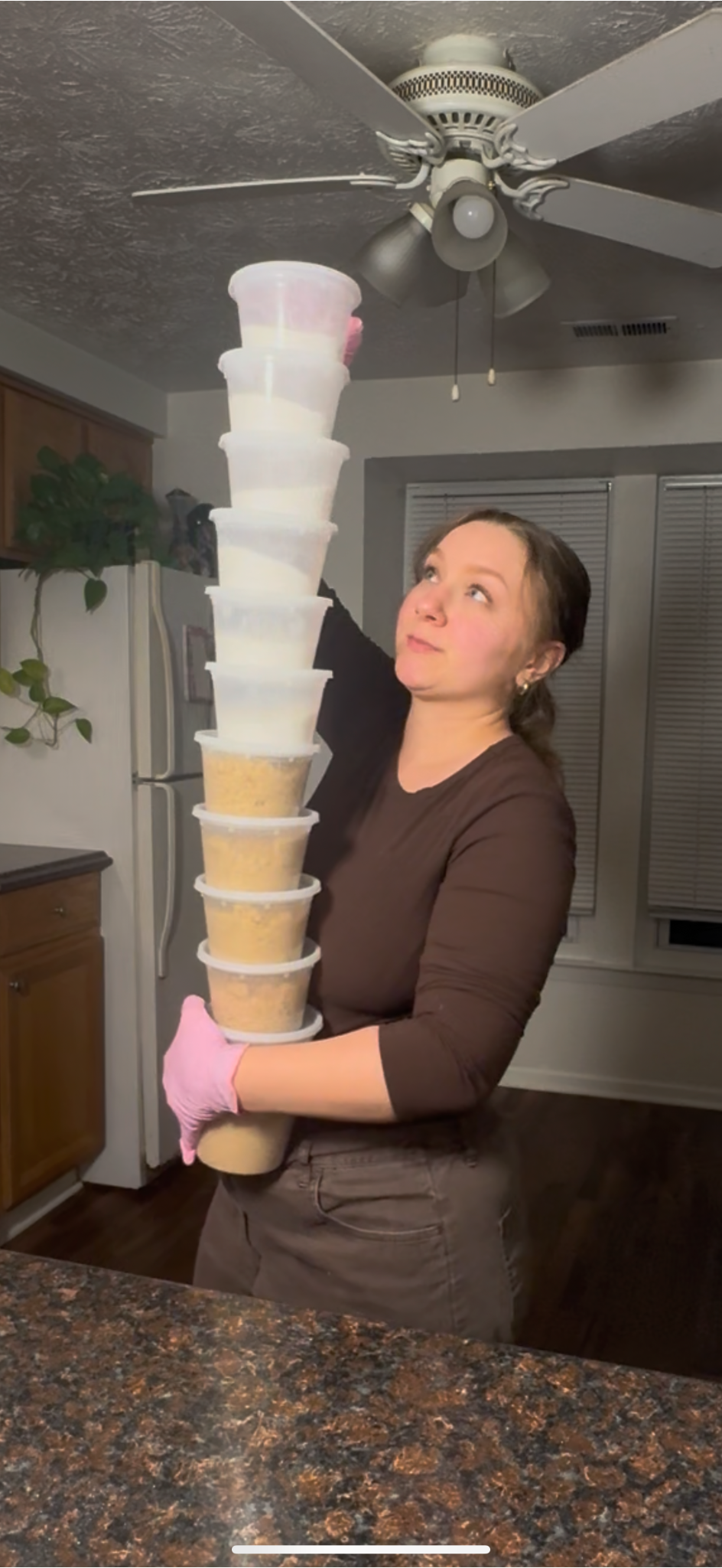 Woman wearing a brown shirt and gray pants holding a tall stack of plastic containers with food inside, in a kitchen.