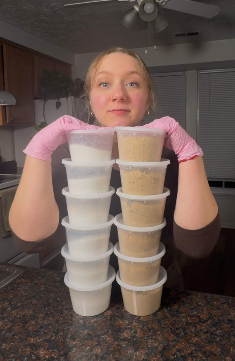 A woman with red hair wearing pink gloves, holding a stack of eight plastic containers filled with white and brown powders, in a kitchen with wooden cabinets and a ceiling fan.