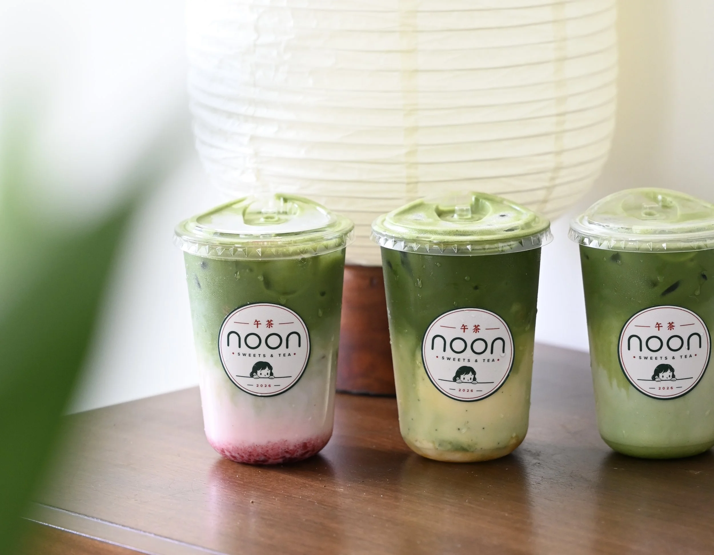 Three plastic cups of matcha-flavored beverages with green tops and a pinkish bottom layer, placed on a wooden surface, with a large paper lantern in the background.