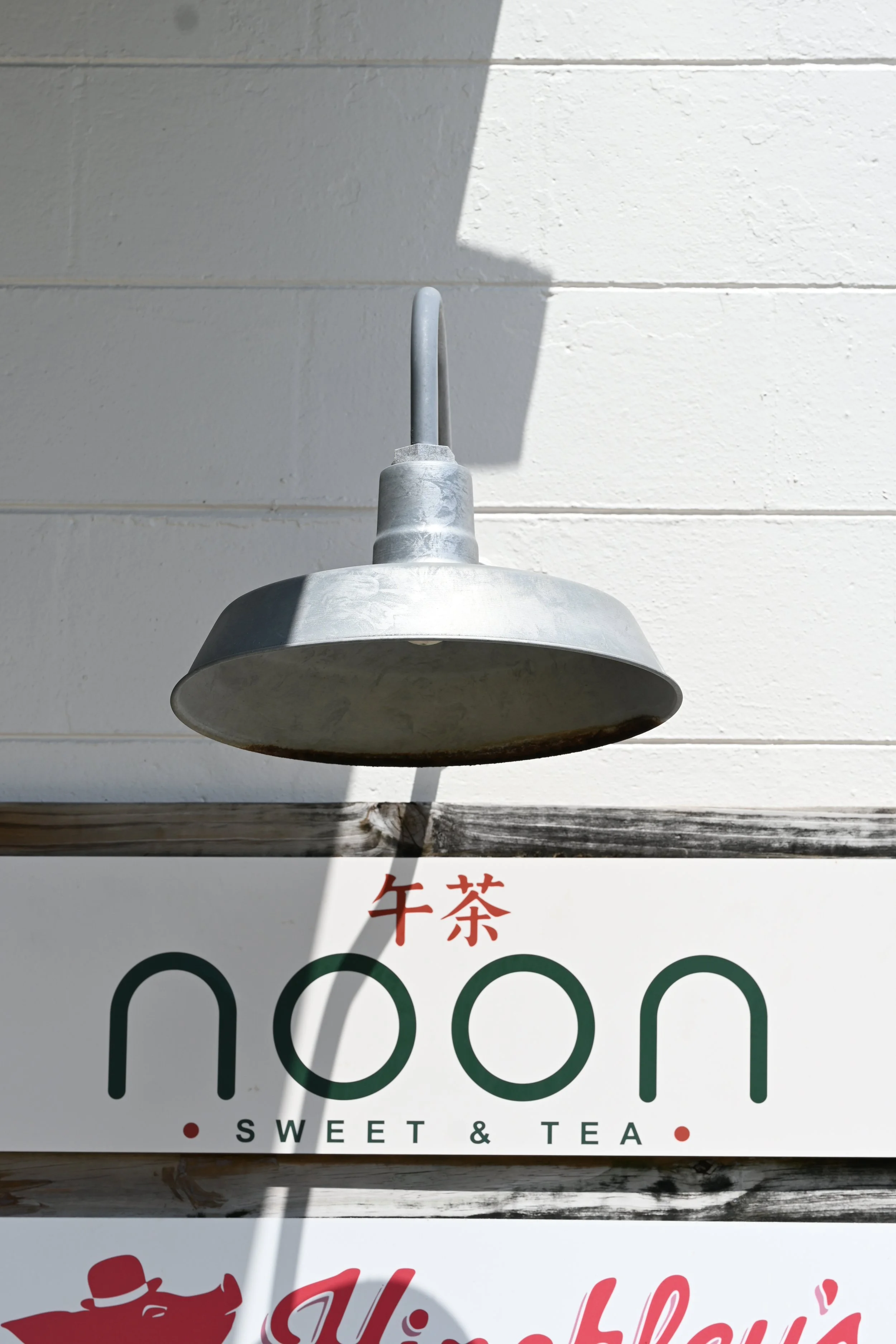 Close-up of a metal outdoor lamp fixture mounted on a white brick wall, with a sign below displaying Chinese characters and the words "Sweet & Tea".