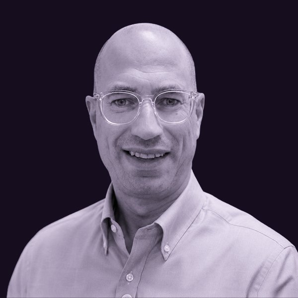 A portrait of Matthew Stremlau smiling man with glasses, wearing a light-colored button-up shirt, against a dark background.
