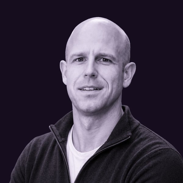 A portrait of Joel Blanchard, wearing a black zip-up jacket and white T-shirt, against a dark background.
