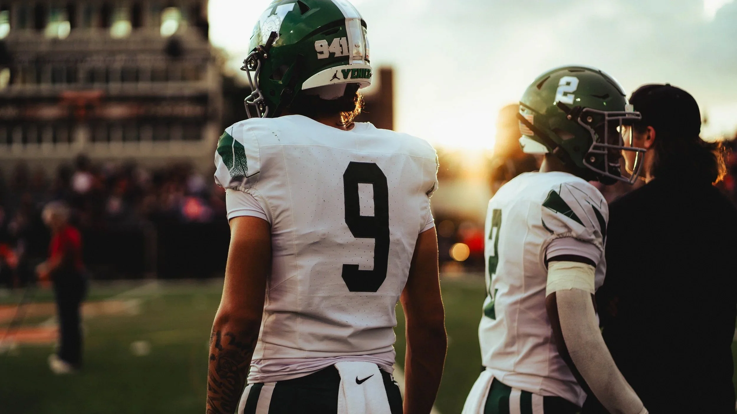 Two football players in white and green uniforms standing on the field during sunset, with one wearing number 9 and the other number 2, while a coach or teammate in black observes in the background.