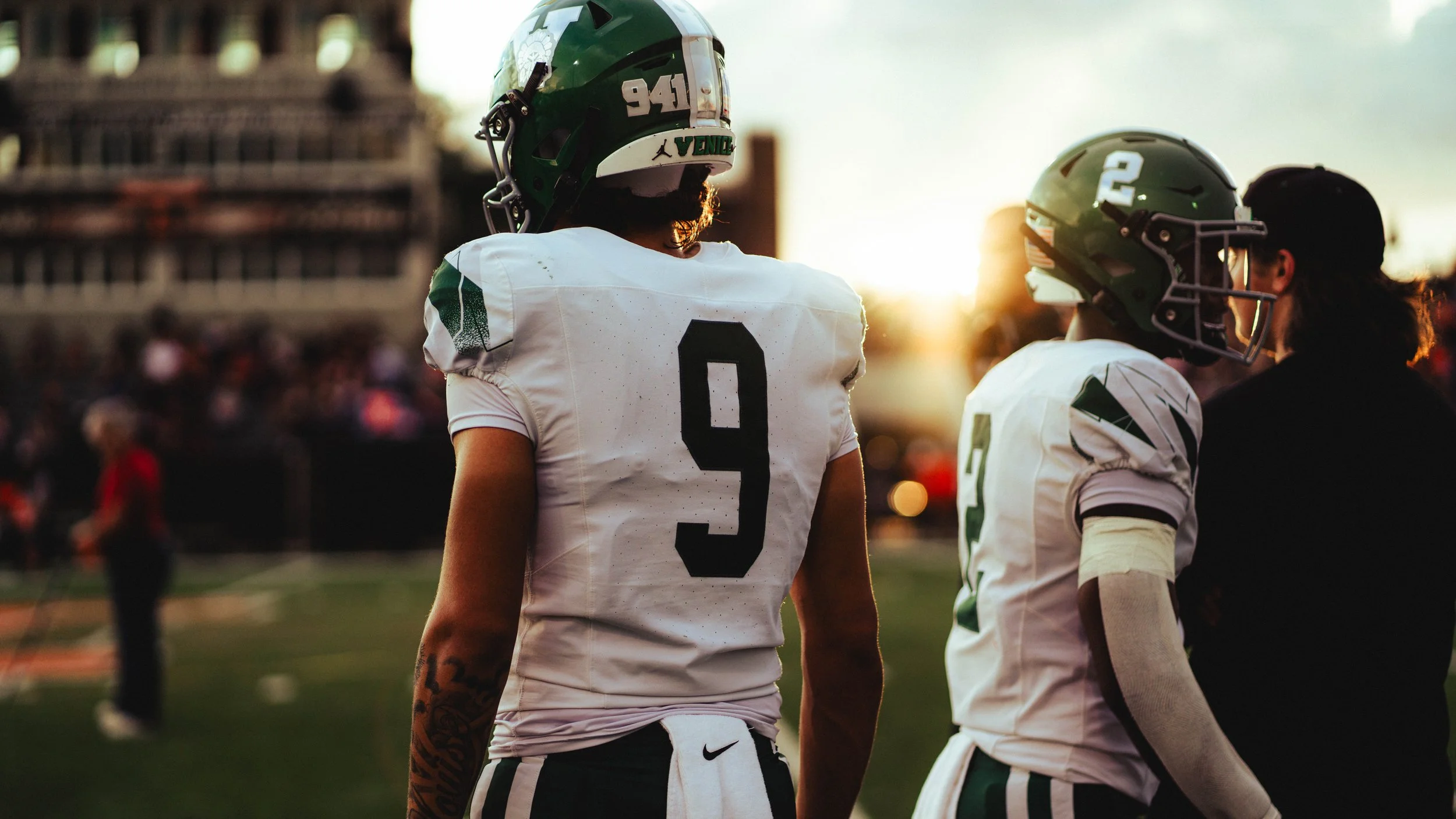 A football player wearing a white jersey with the number 9 and a green helmet stands on a football field during sunset, with two other players visible in the background.