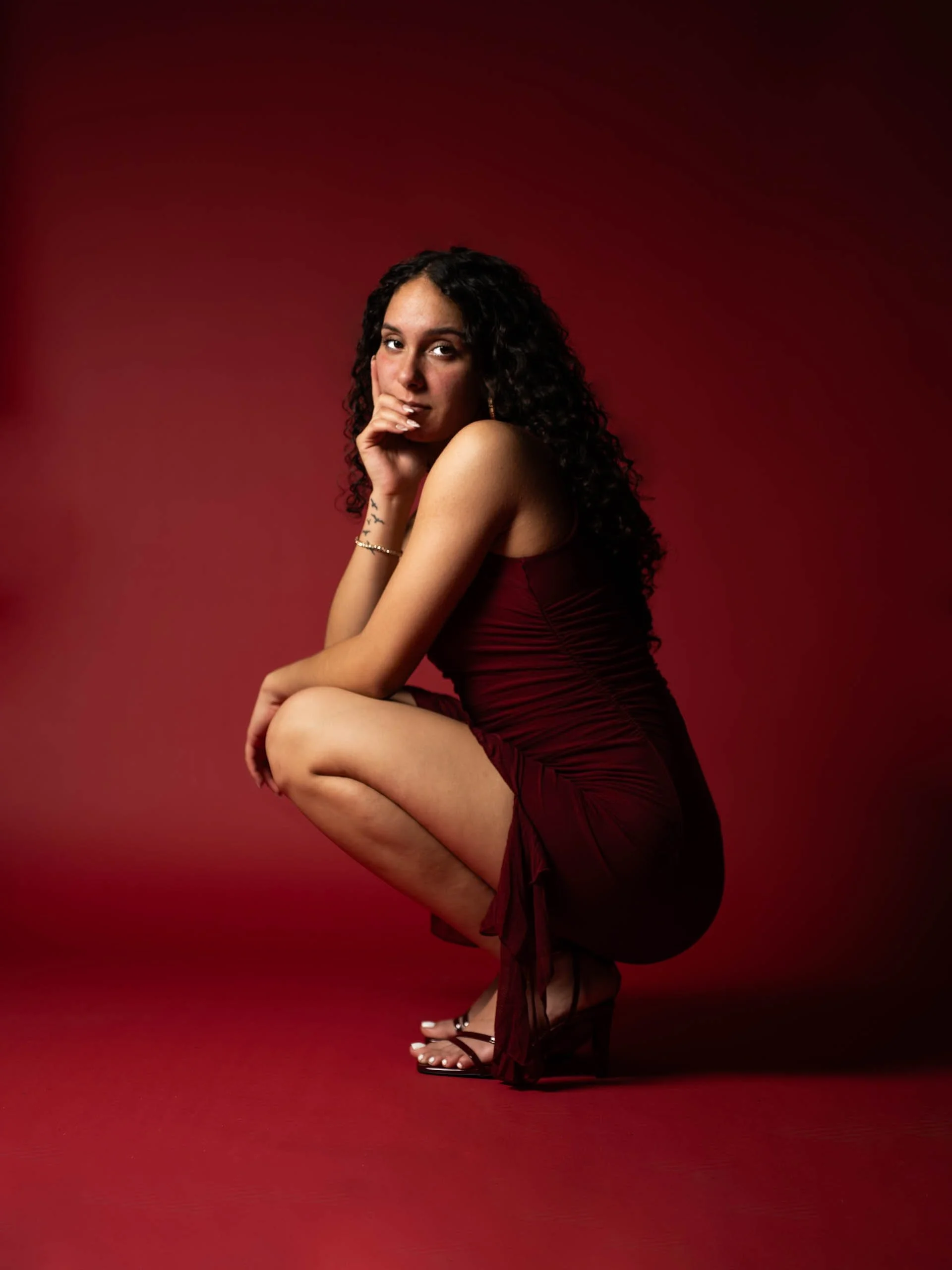 A woman with curly dark hair, wearing a red dress, squatting and looking at the camera with a thoughtful expression, against a red background.