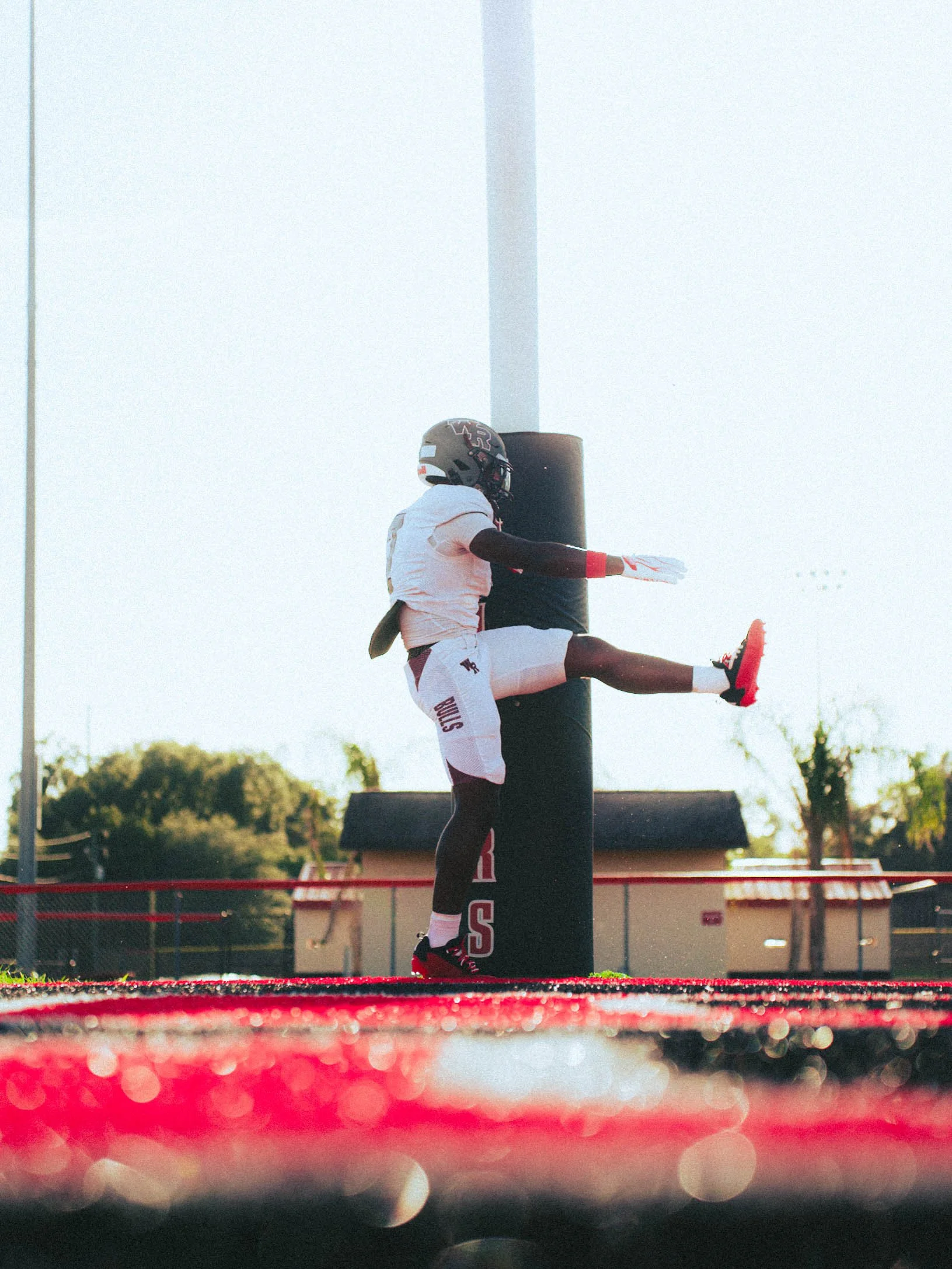 A football player in a white uniform with red and black accents is performing a high kick on a football field, with a black padded goalpost behind him and trees and a building in the background.