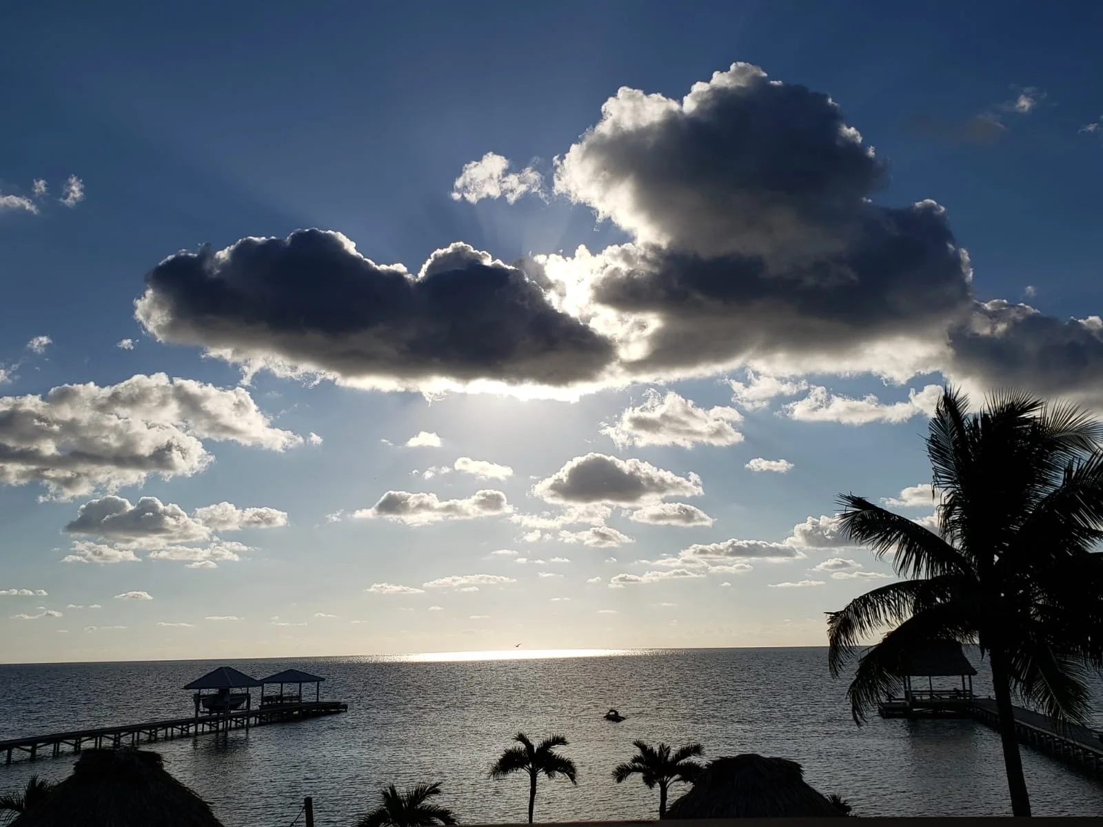 Sunset over the ocean with a partly cloudy sky, palm trees, and a wooden pier extending into the water.