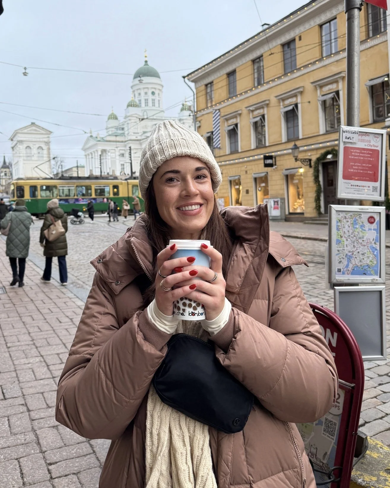 A woman with dark hair wearing a beige knitted beanie and a brown puffer jacket, holding a cup of coffee and smiling, standing in a city square with historic buildings and a tram in the background.