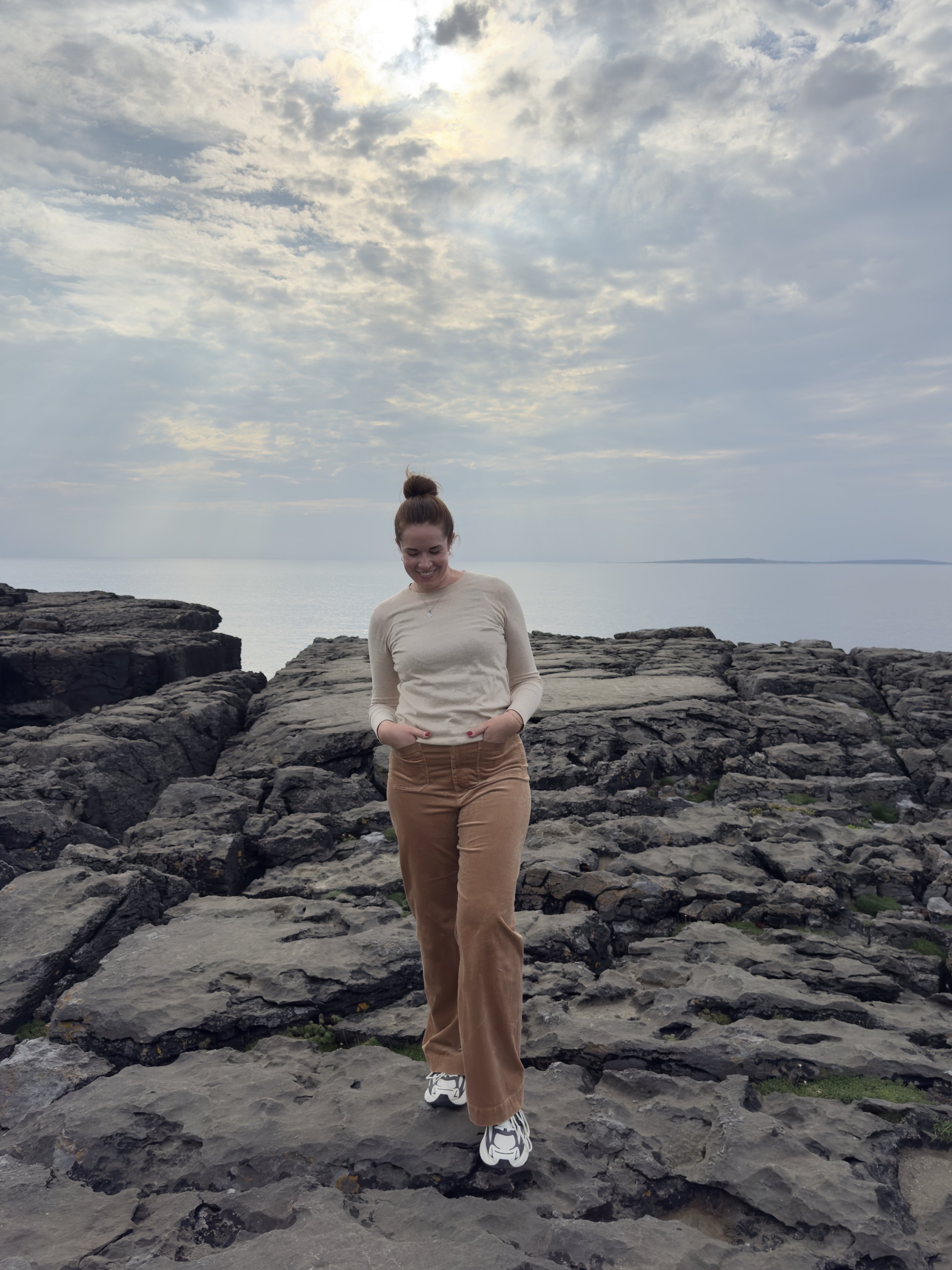 A young woman with a bun hairstyle standing on rocky terrain near water, with cloudy sky overhead and the sun partially obscured by clouds.