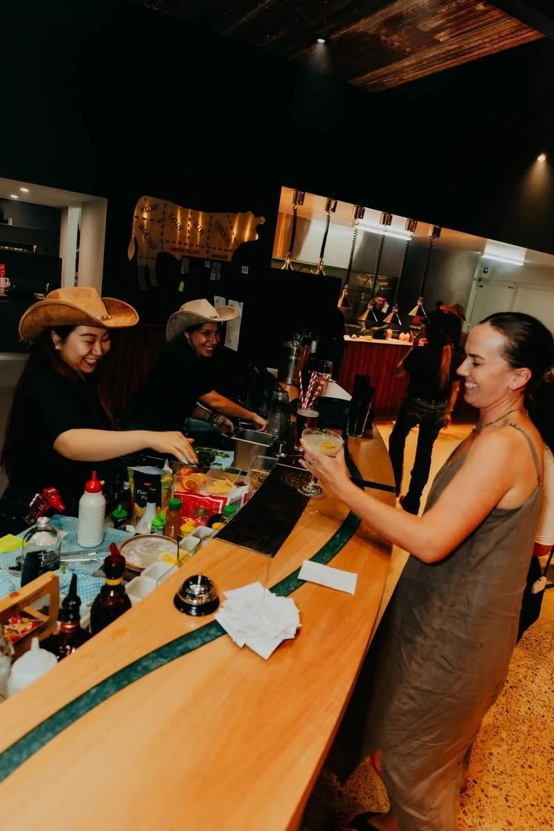 Bluehole Steakhouse bar in service, with staff pouring drinks across the curved timber bartop and its blue resin river. View the Bluehole Bar project.