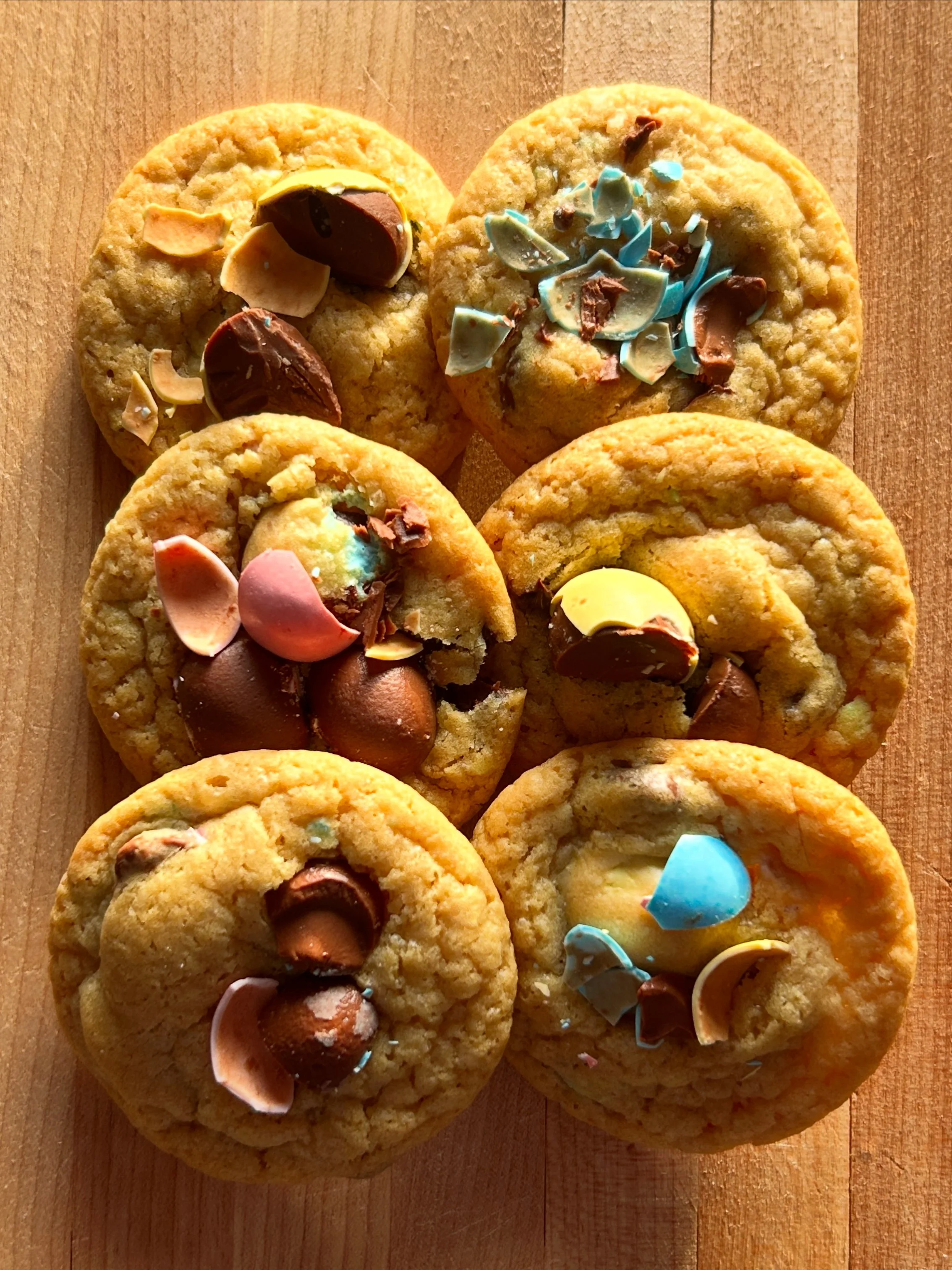 Six cookies topped with chocolate, colorful sprinkles, and broken chocolate pieces arranged on a wooden surface.