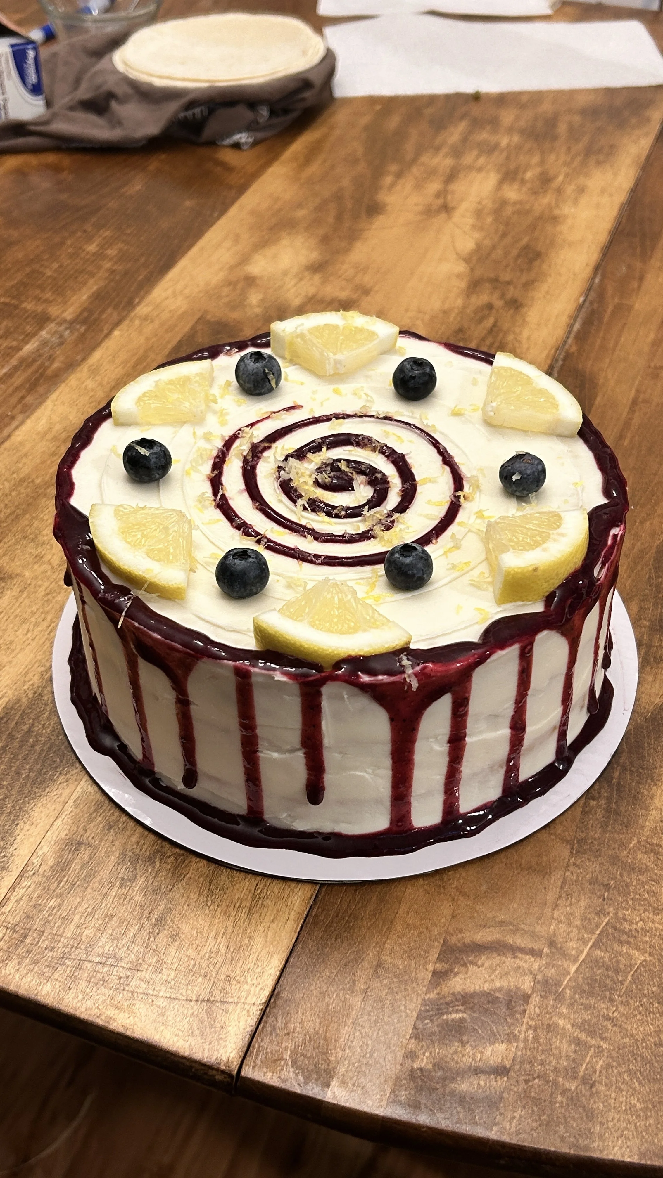 A round cake with white frosting, decorated with lemon wedges and blueberries on top, and red chocolate drip around the sides, on a wooden table.