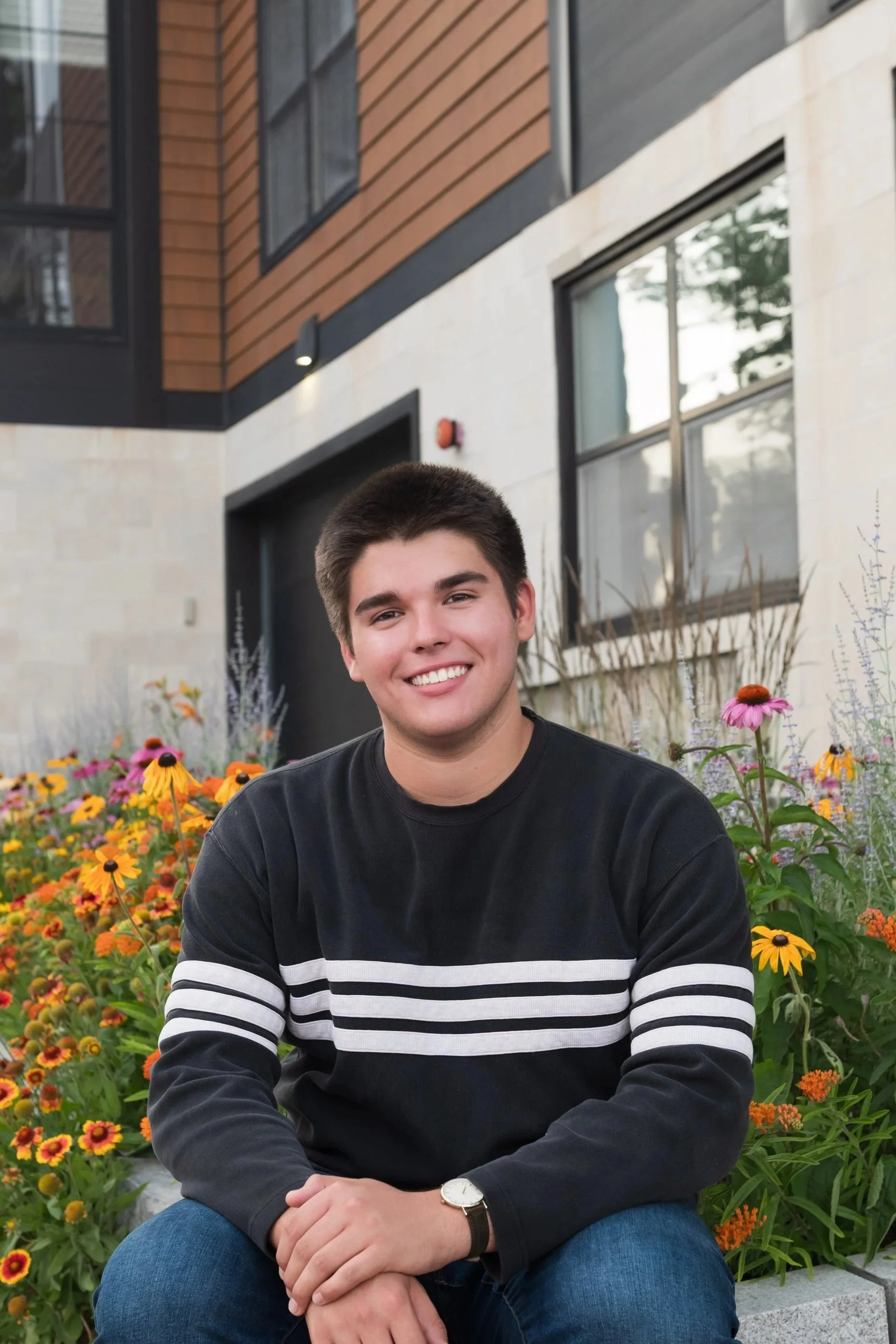 A young man with short dark hair, wearing a black and white striped sweater and a watch, sitting in front of colorful flowers outside a modern building with large windows and wooden paneling.