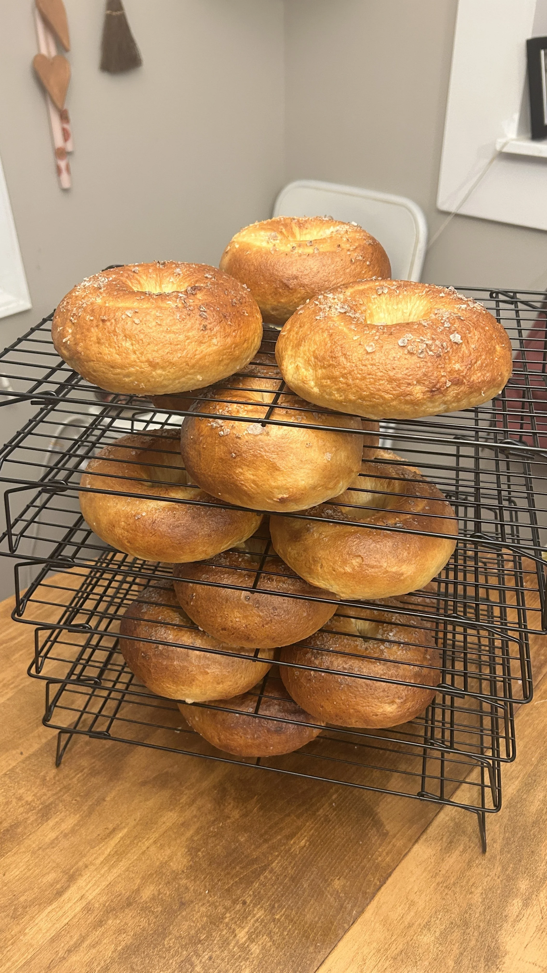 Several freshly baked bagels with a golden-brown crust and coarse salt on top, stacked on three-tier wire racks on a wooden table.
