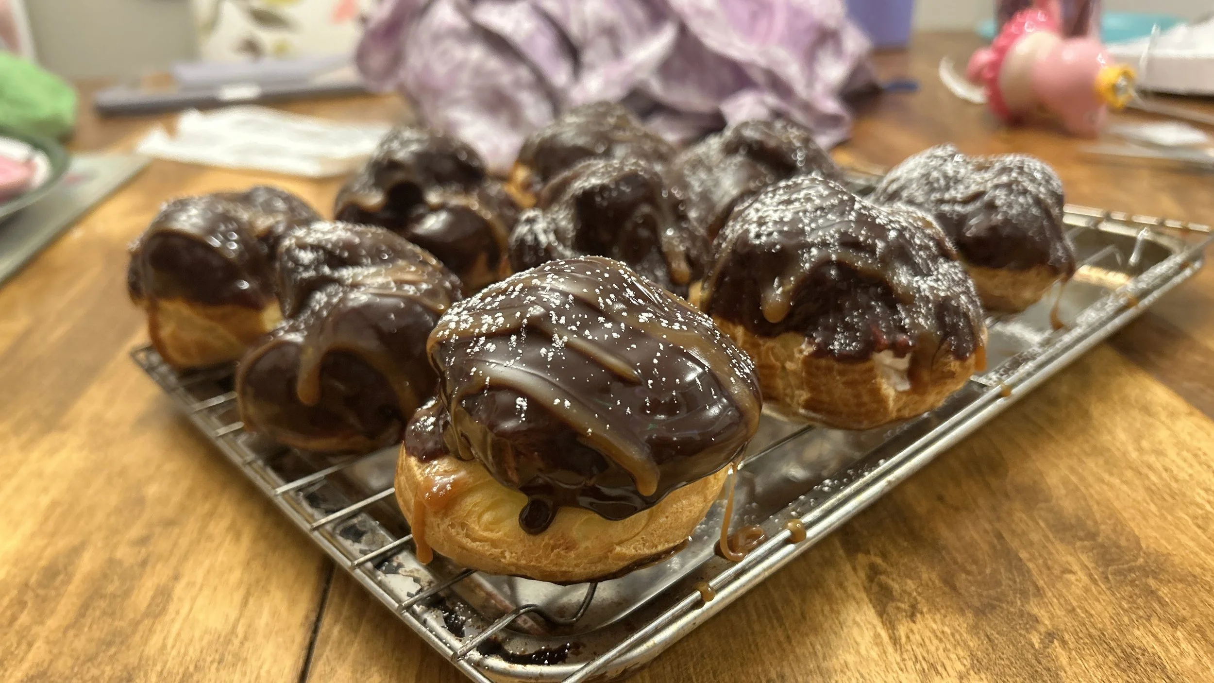 A tray of cream puffs topped with chocolate glaze and drizzled with caramel sauce on a wooden table.