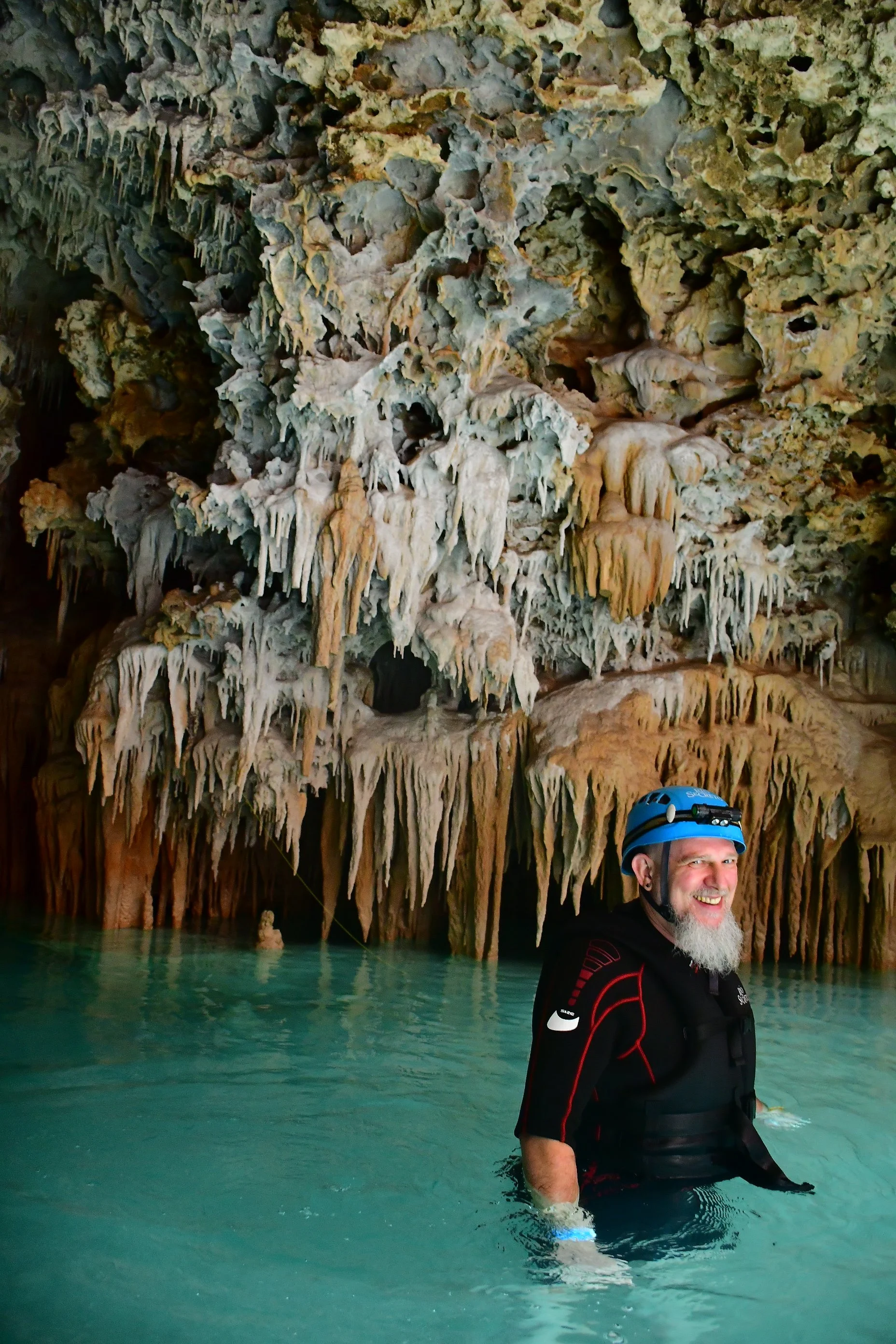 The founder wearing a helmet and a wetsuit stands in clear green water inside a cave or cavern with stalactites and stalagmites, smiling at the camera.