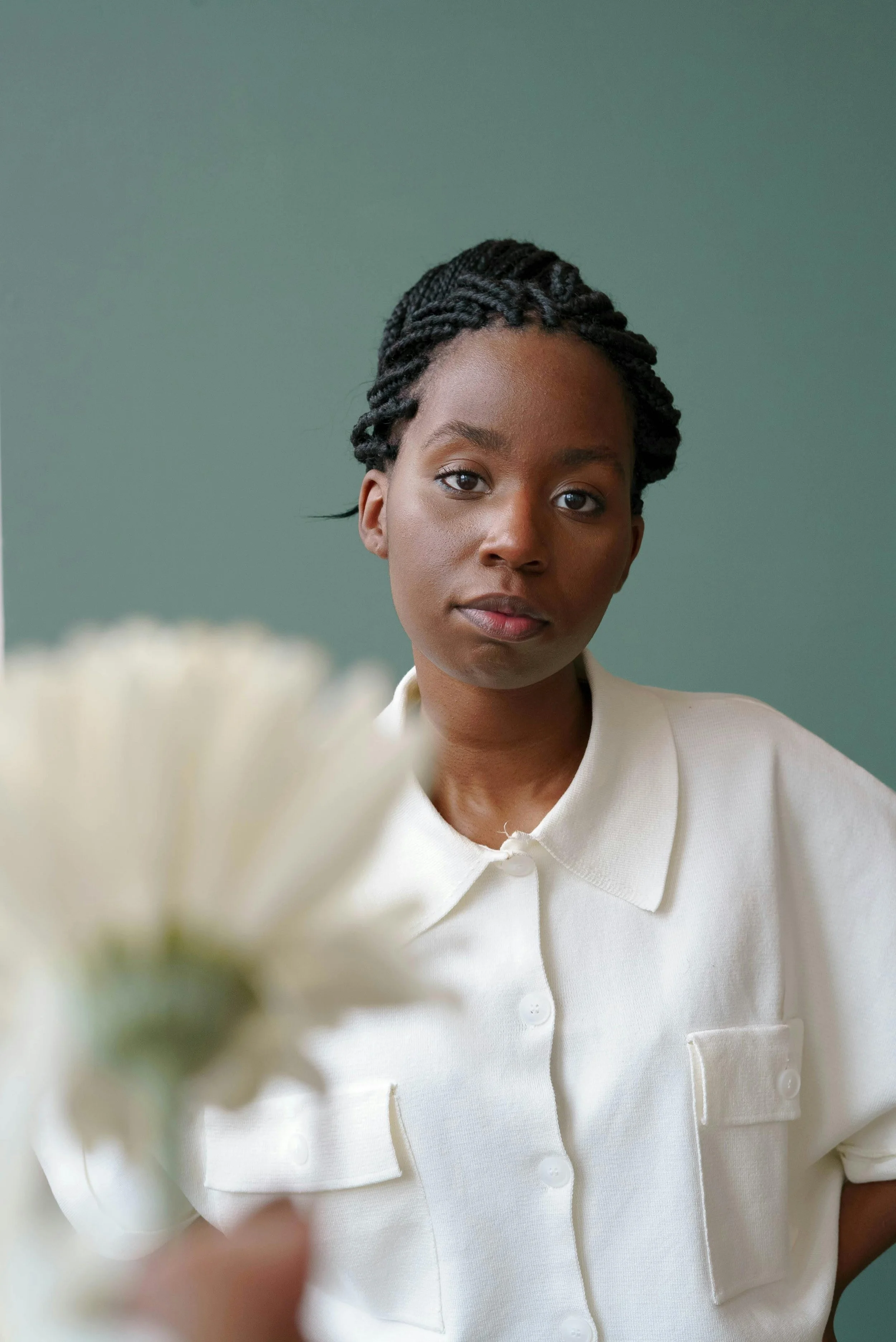 A close-up image of a woman with dark skin and braided hair, wearing a white collared shirt, looking at the camera against a muted green background.
