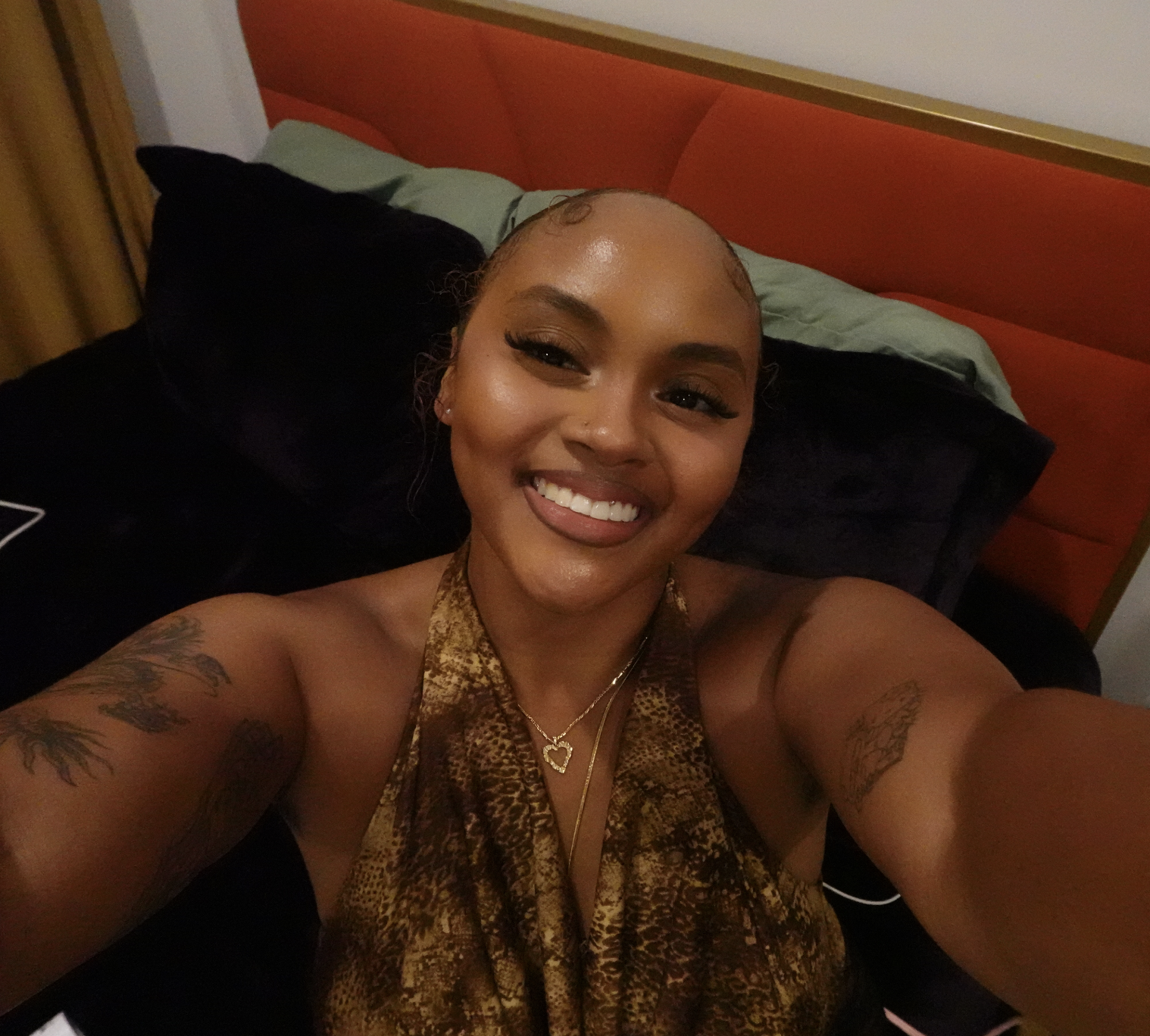 A woman with tattoos on her arms, smiling and taking a selfie in a room with a red headboard, pillows, and a yellow curtain.
