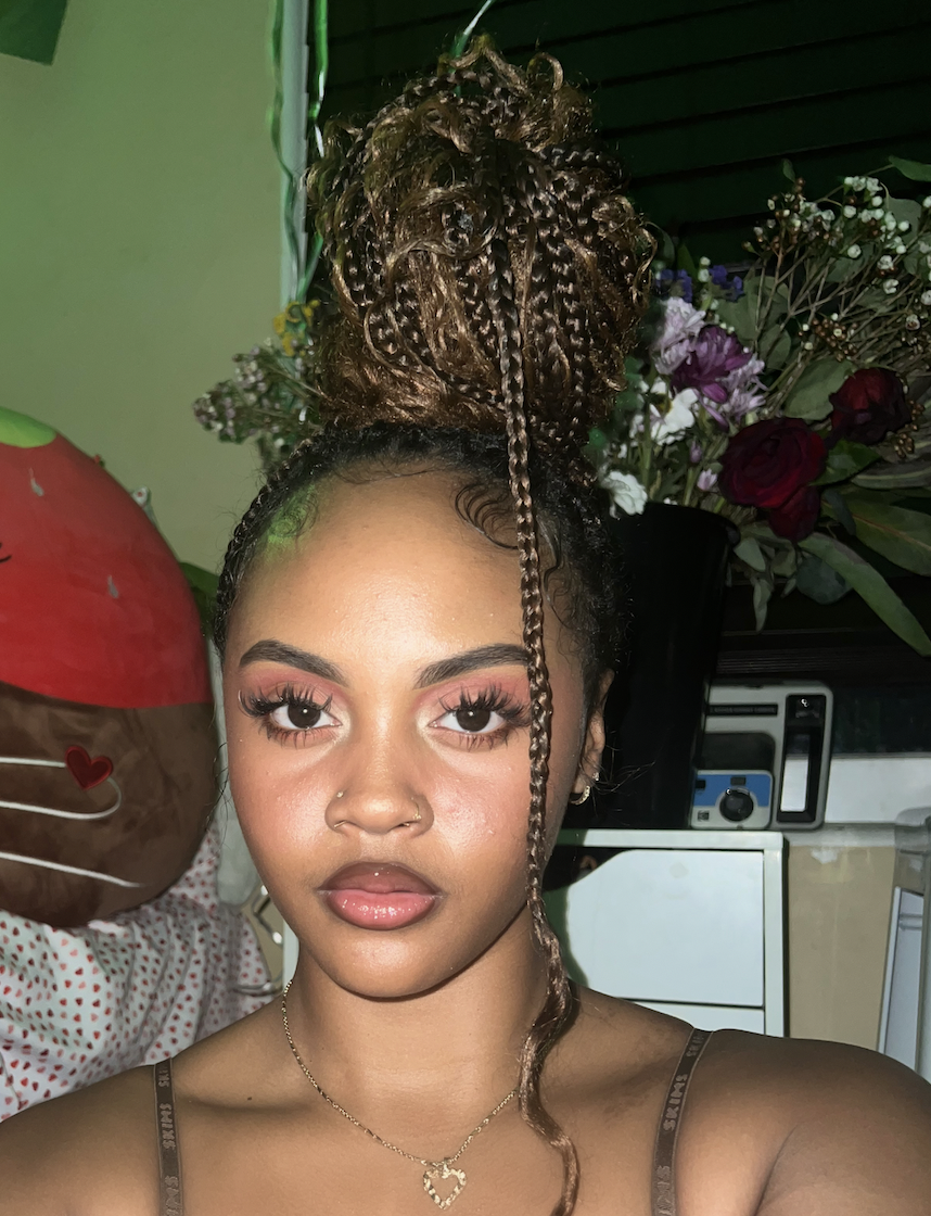 Young woman with styled curly hair in a high bun with braid, wearing makeup, a nose ring, and a heart-shaped necklace. Background includes flowers and some electronic equipment.