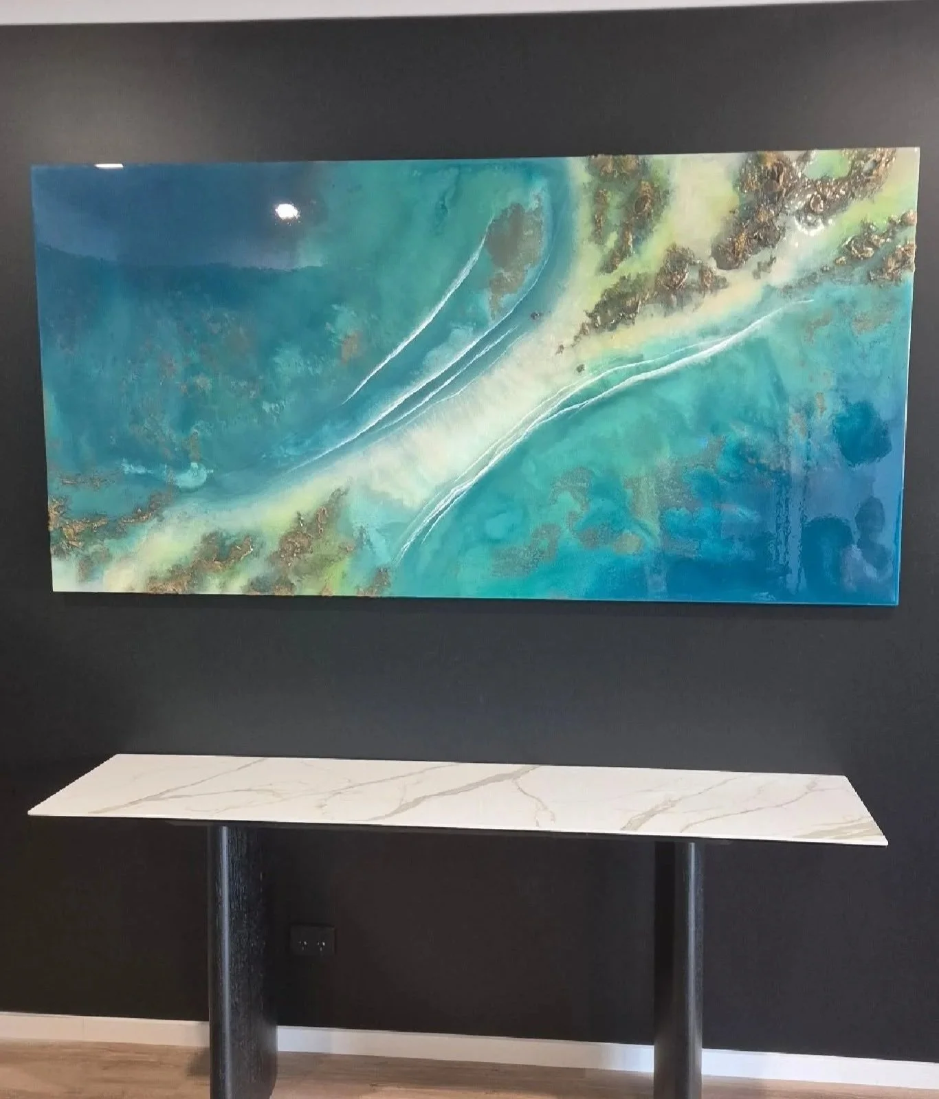 Abstract watercolor painting with blue, green, and gold tones hung on a black wall, displayed above a white marble table with black metal legs.