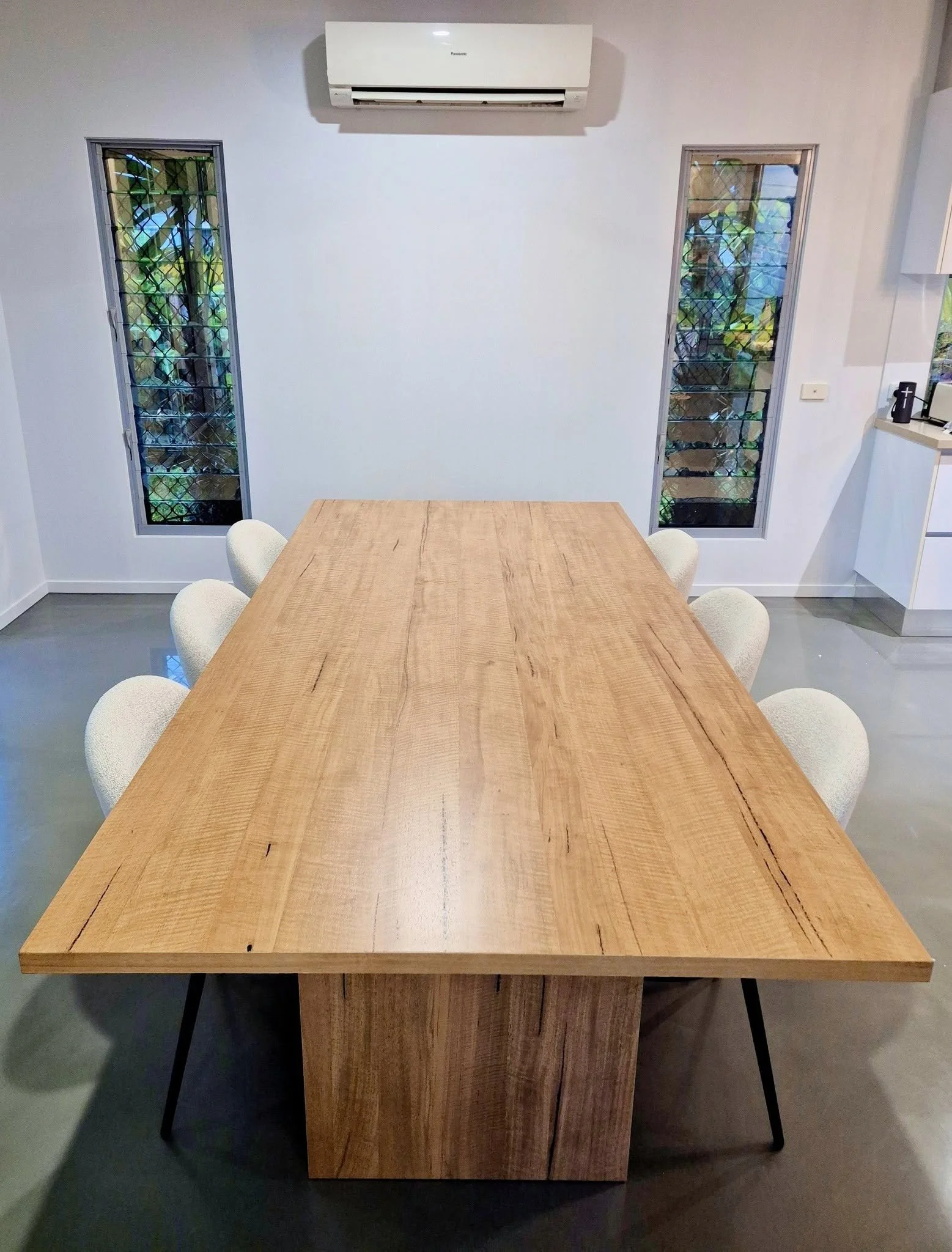 A wooden dining table with six beige upholstered chairs in a room with white walls, two narrow windows with metal grilles, a white wall-mounted air conditioner, and a small kitchen area with a black kettle and toaster in the background.