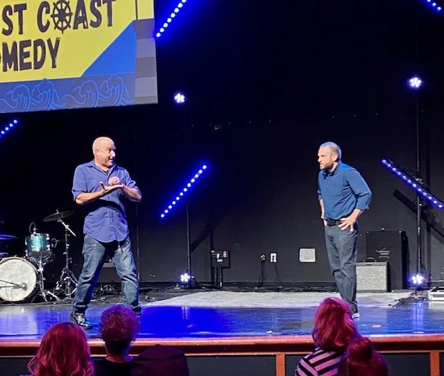 Two men stand on stage in front of an audience at a comedy show. One man is speaking, gesturing with his hands, while the other listens. Stage lights and a drum set are visible in the background, with a sign that reads 'West Coast Comedy' partially visible at the top.