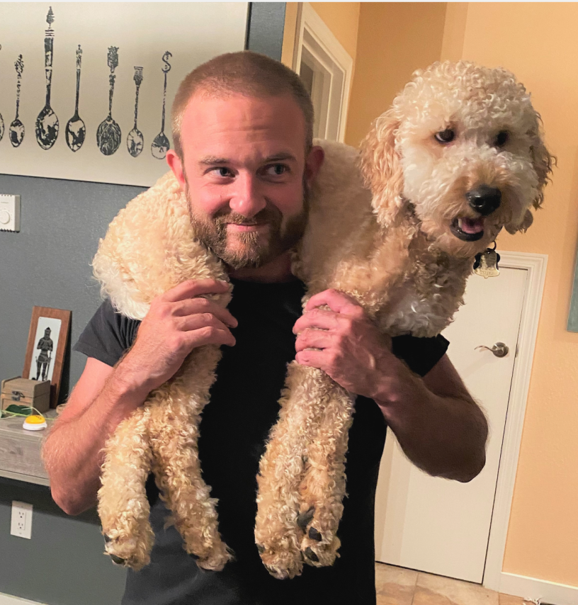 A man with a beard holding a curly-haired dog over his shoulders inside a house.