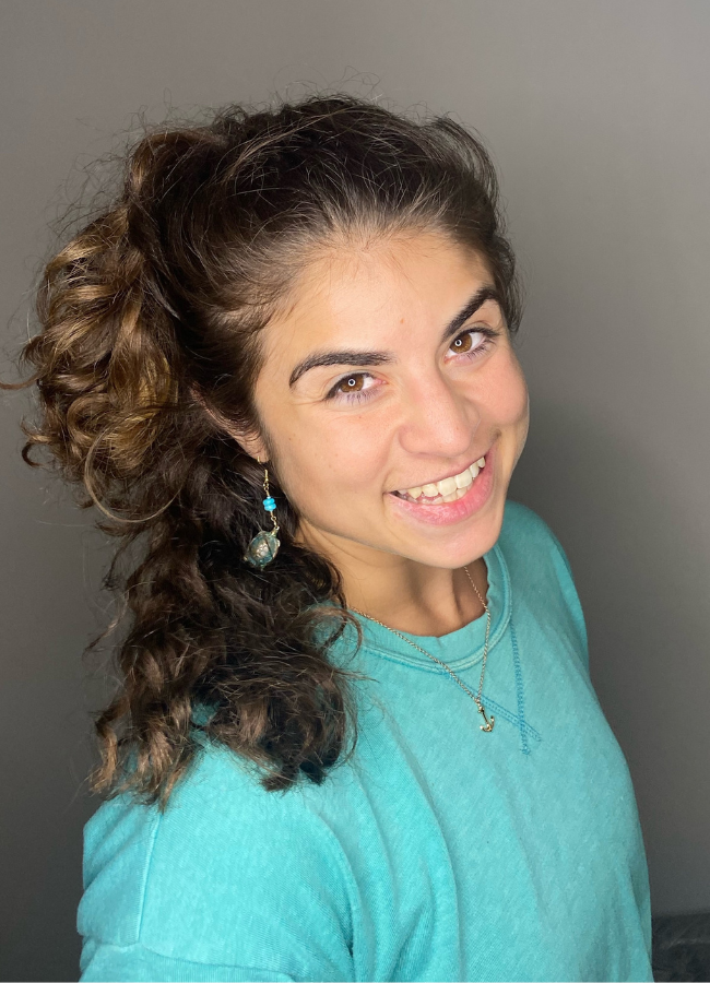 Young woman with curly brown hair and light skin smiling, wearing a turquoise shirt, a necklace, and a dangling earring, against a neutral background.