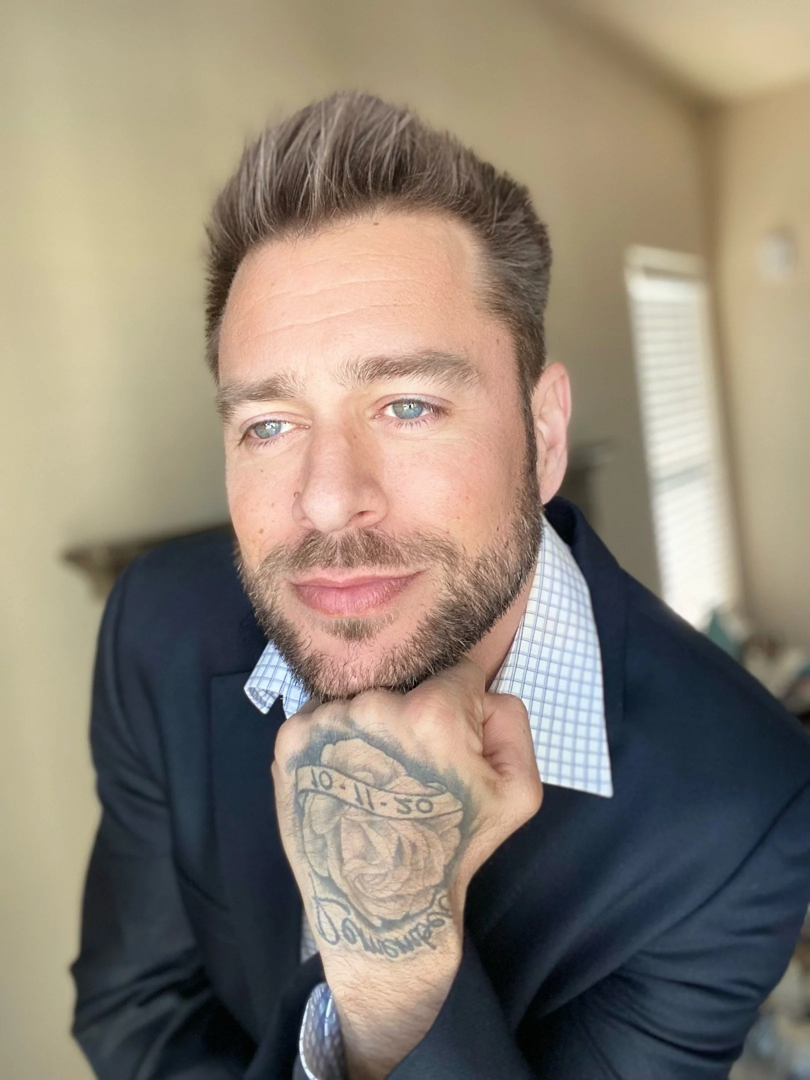 A close-up portrait of a man with blue eyes, a beard, and styled hair, wearing a dark suit and light checkered shirt, resting his chin on his hand with a tattooed tattooed wrist, in an indoor setting with a blurred background.