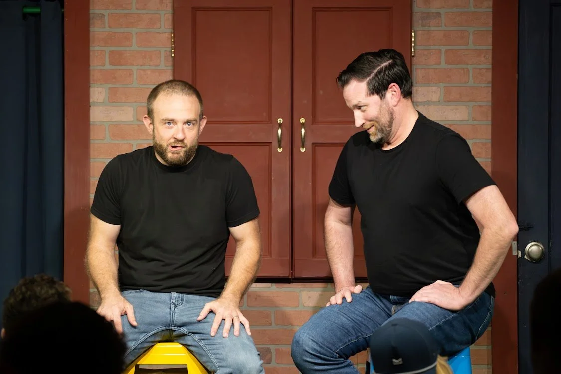 Two men sit on stools on a stage with a brick wall background, wearing black t-shirts and jeans, engaged in a conversation.