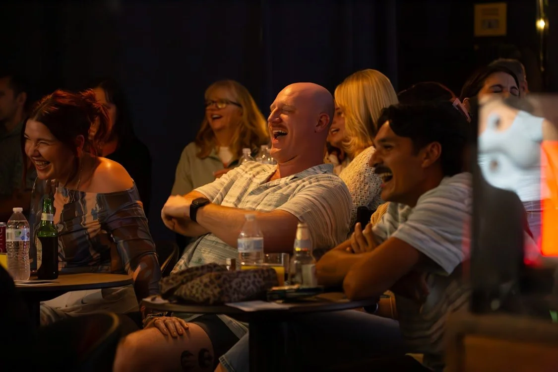 A group of people sitting at a table, laughing and enjoying a comedy show or performance in a dimly lit venue.