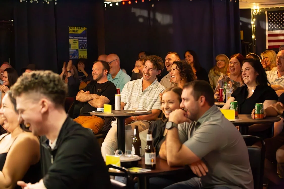 Audience members at a comedy show, smiling and laughing.