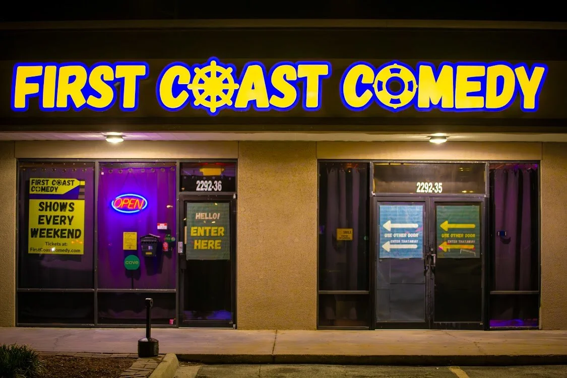 Exterior of a comedy club named First Coast Comedy at night, featuring a neon 'Open' sign, signs instructing entry, and promotional posters indicating shows every weekend.
