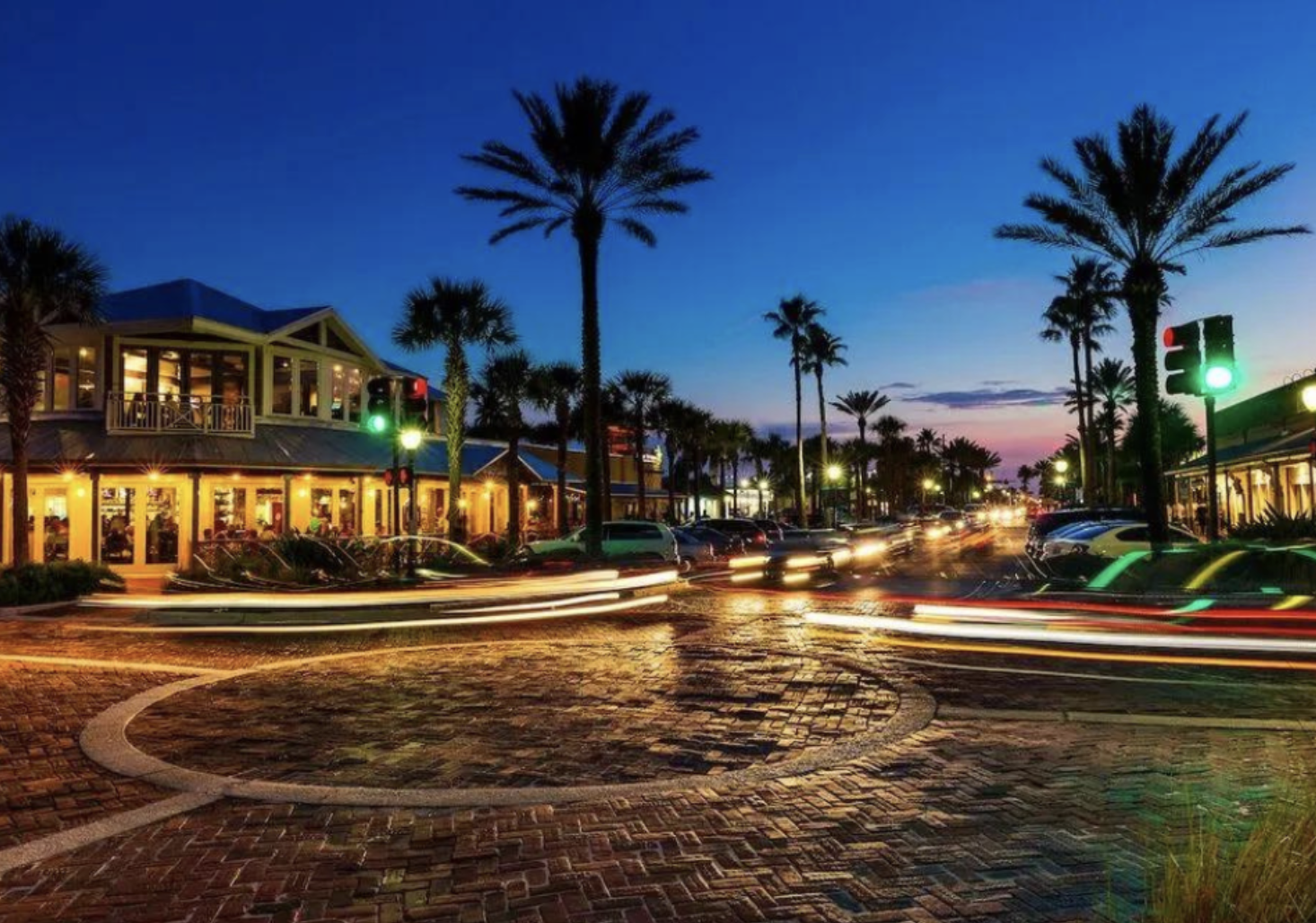 A lively street scene at dusk with palm trees, illuminated shops, and a restaurant with large windows on the left. Cars are moving through the intersection, leaving light trails, and traffic lights are green. The sky is a gradient of blue and pink, and streetlights cast a warm glow.