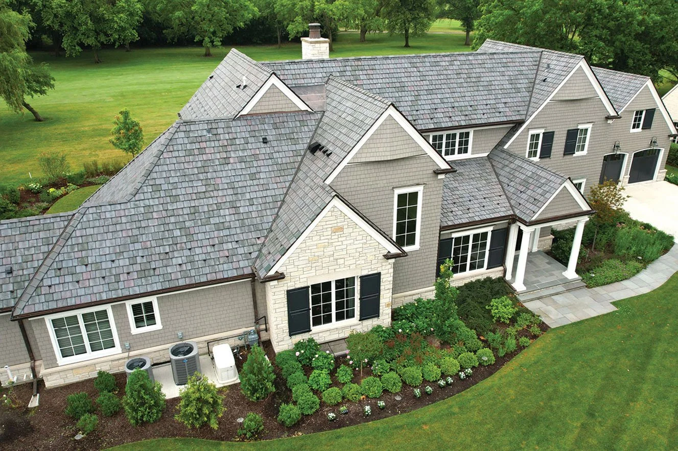 A large house with multiple gabled roofs, gray siding, and white accents, surrounded by a well-maintained lawn and landscaped garden with bushes and trees.