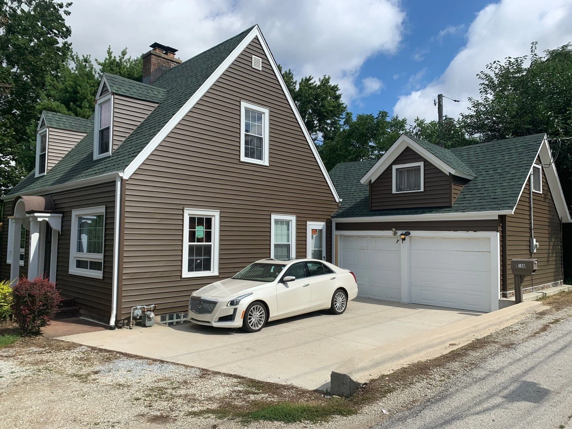 A two-story house with beige siding, a dark gray gable roof, and white trim. The house has multiple windows, a garage door, and a white car parked in the driveway. There are trees and a partly cloudy sky in the background.