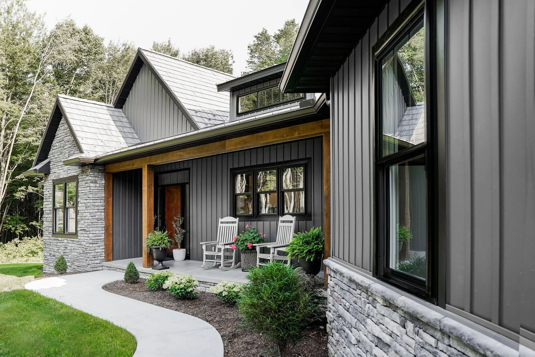 Modern house with black siding, stone accents, and large windows, featuring a backyard patio with white chairs, potted plants, and a curved concrete walkway, surrounded by green lawn and trees.