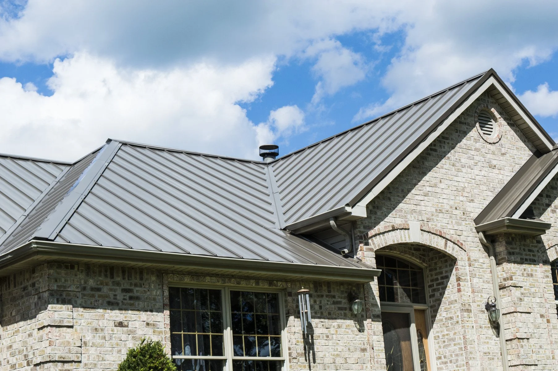 Close-up of a house with a metal roof, brick exterior walls, a chimney, and a large window under a blue sky with clouds.