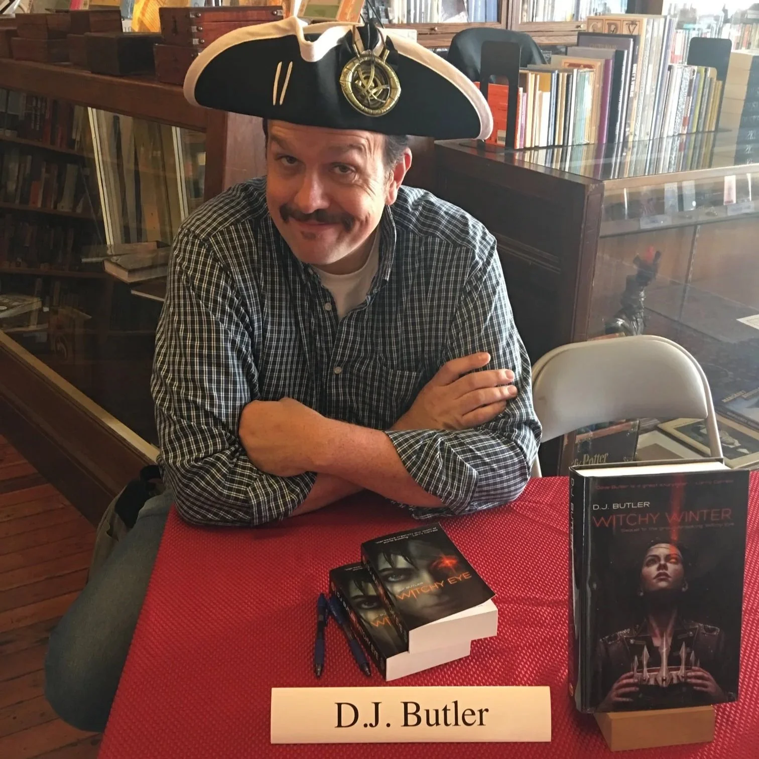 A man with a mustache and beard wearing a checkered shirt and a pirate hat with a golden emblem inside an indoor library or bookstore. He's sitting at a table with a red tablecloth, surrounded by books and promotional materials for the book titled "Witchy Winter" by D.J. Butler. There is a name tag on the table reading "D.J. Butler."