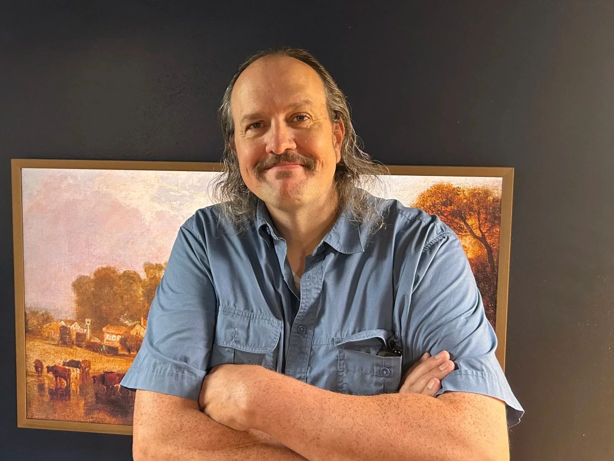 A middle-aged man with long brown hair and mustache, wearing a gray button-up shirt, sitting with his arms crossed in front of a framed landscape painting of a rural scene with trees and fields.