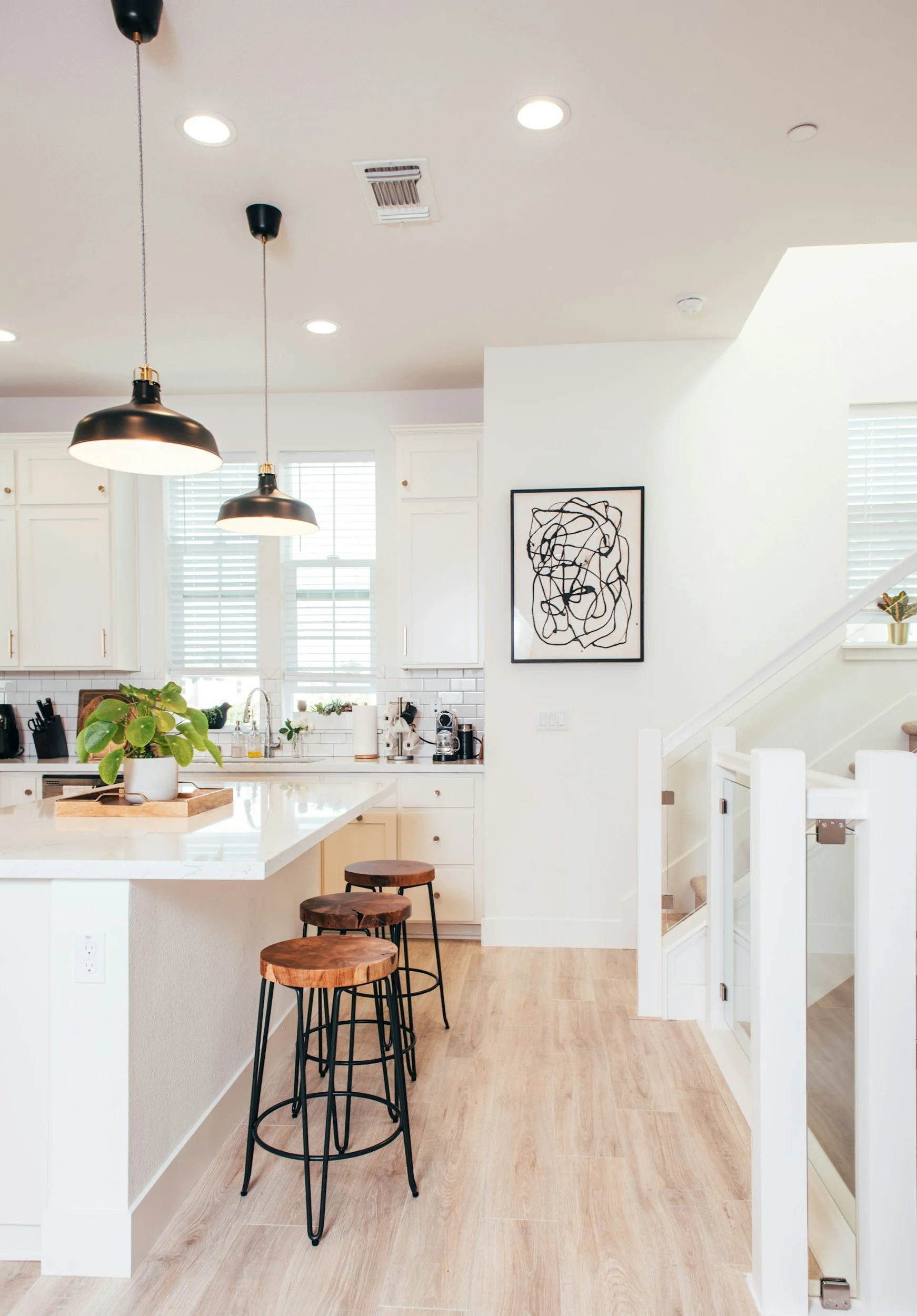 Modern kitchen with white cabinets, black pendant lights, artwork on the wall, a white island with wooden stools, and large windows with blinds.