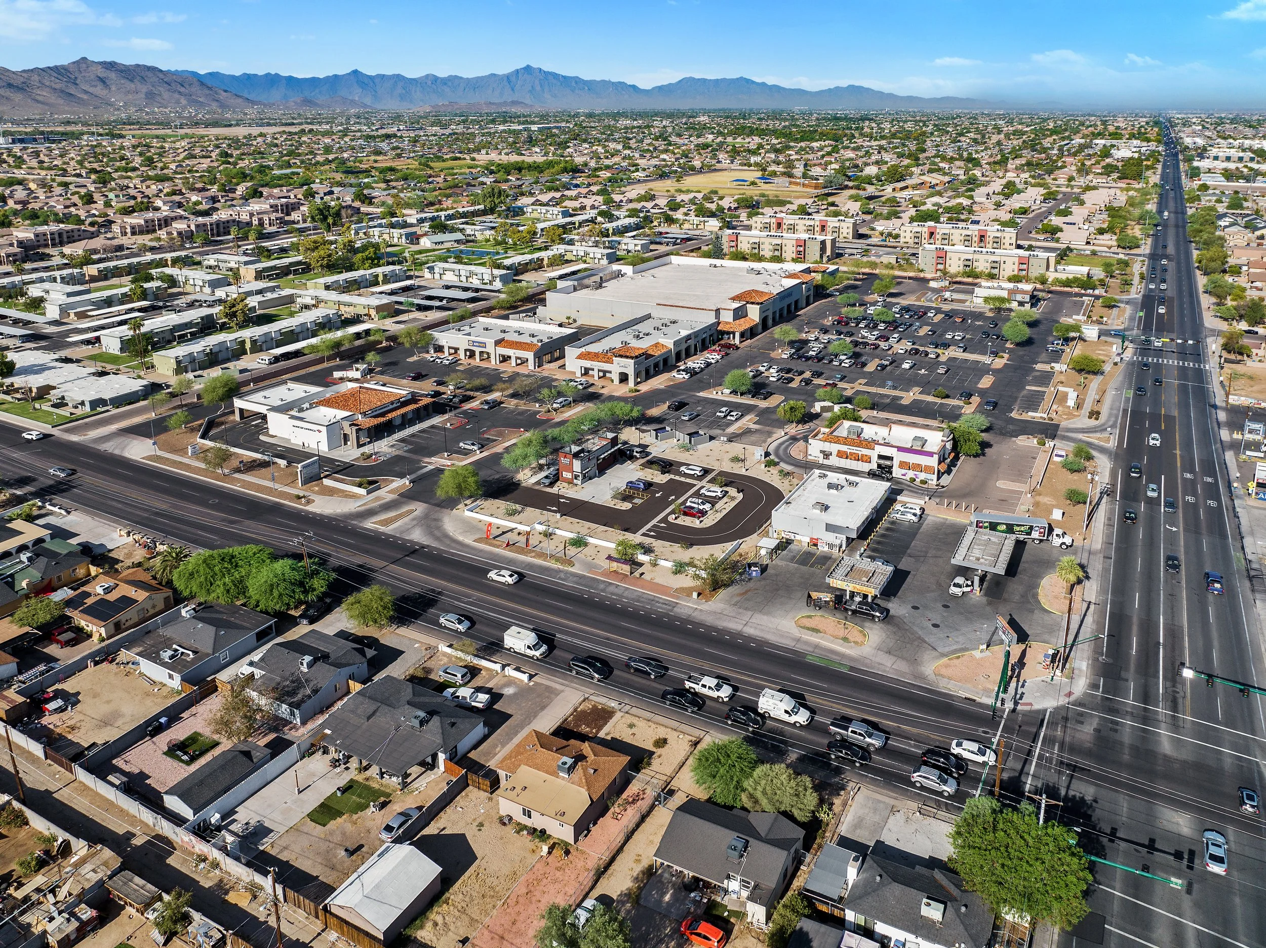 An aerial view of a suburban area with houses, apartments, a shopping center, and a main road extending into the distance, with mountains in the background under a clear blue sky.