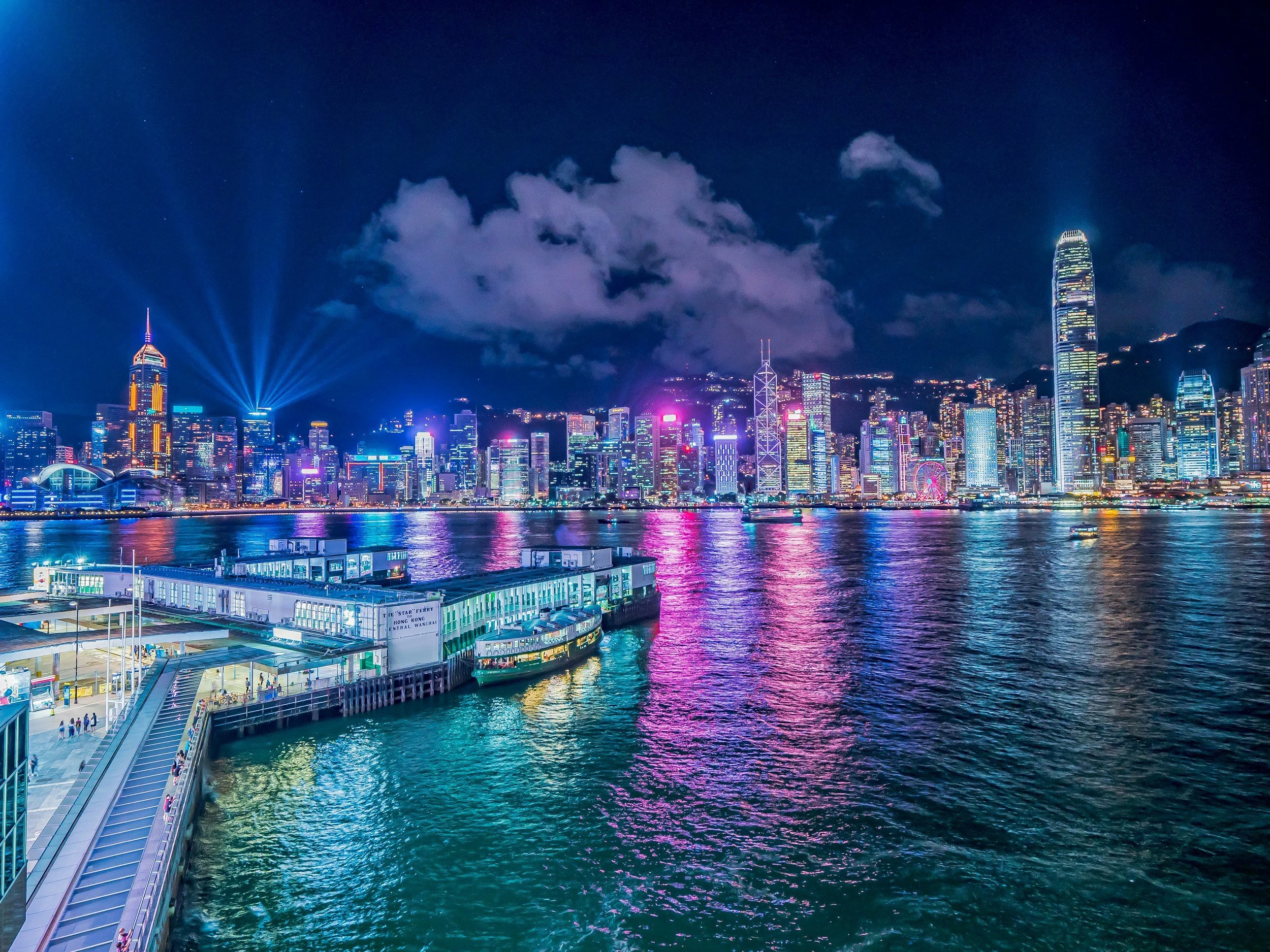 Hong Kong skyline at night with colorful lights, tall buildings, and reflections on the water, including a river ferry terminal in the foreground.